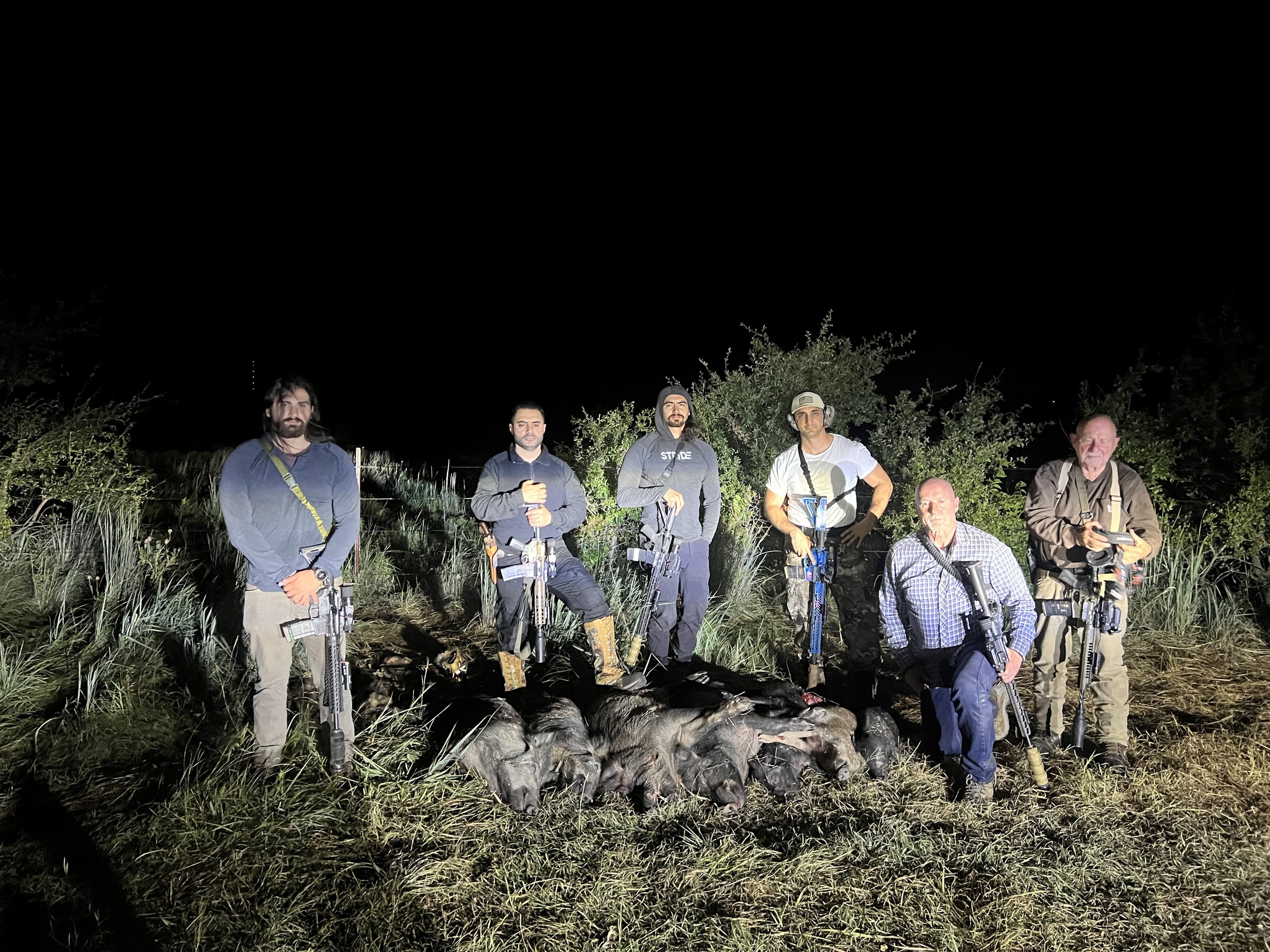 Group of six men standing outdoors at night, some holding rifles, with a large pile of what appears to be animal carcasses in front of them, surrounded by tall grass and bushes.