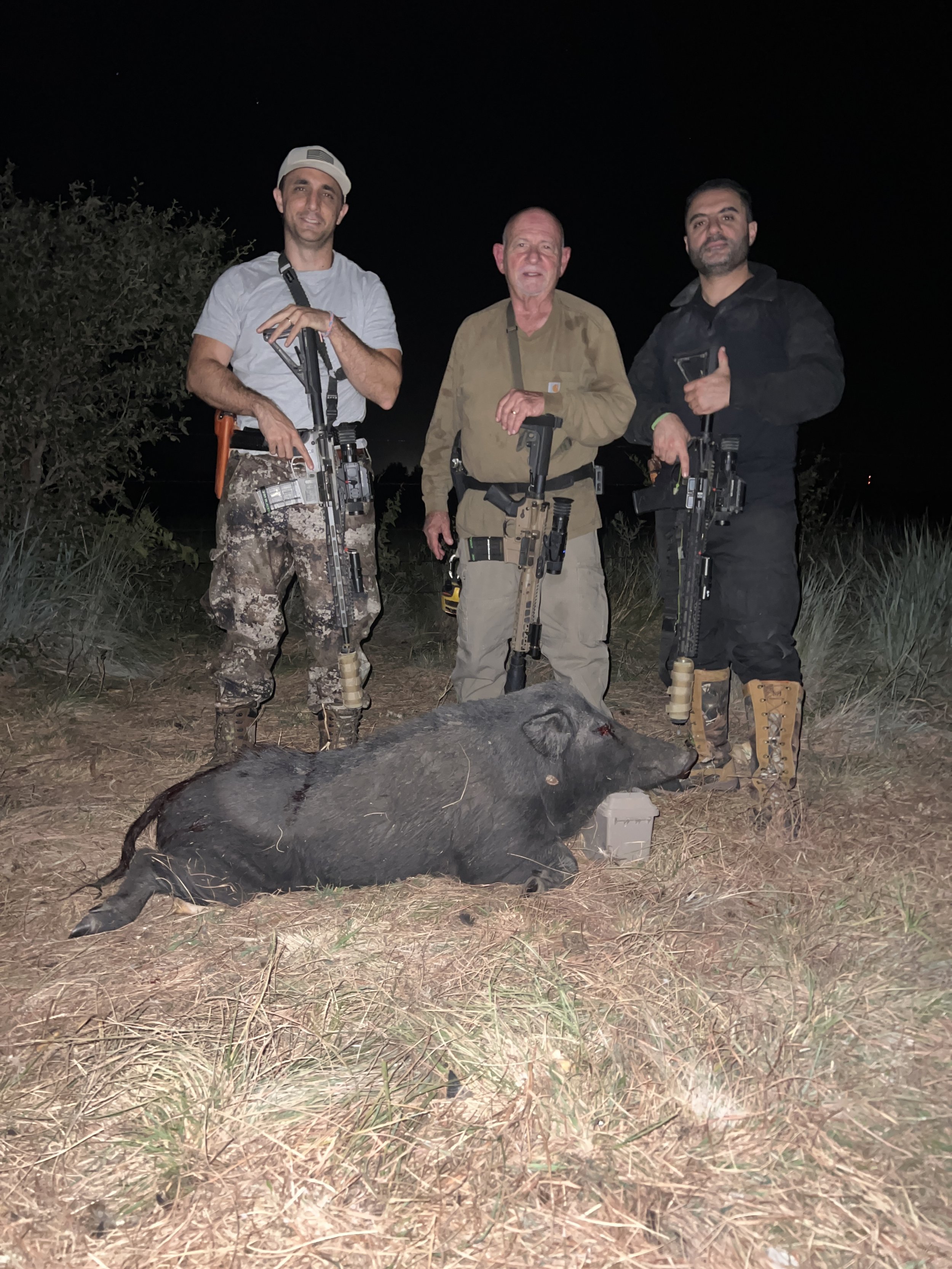 Three men standing outdoors at night with rifles, posing behind a large dead boar on the ground, surrounded by grass and bushes.