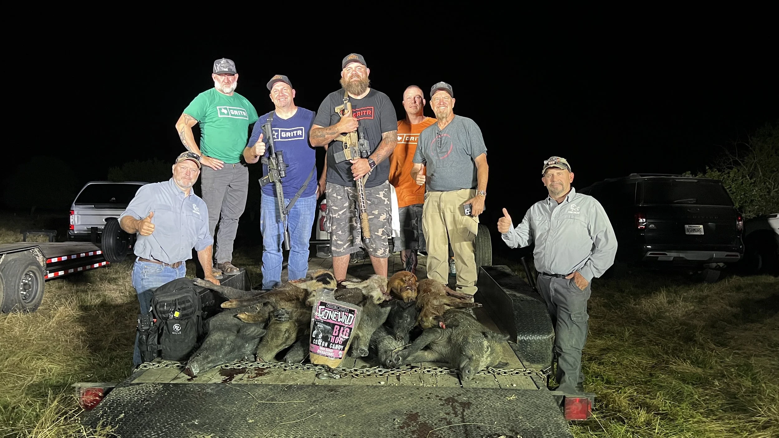 A group of men posing outdoors at night with hunting dogs and a display of hunted animals, likely after a hunting trip, with greenery and vehicles in the background.