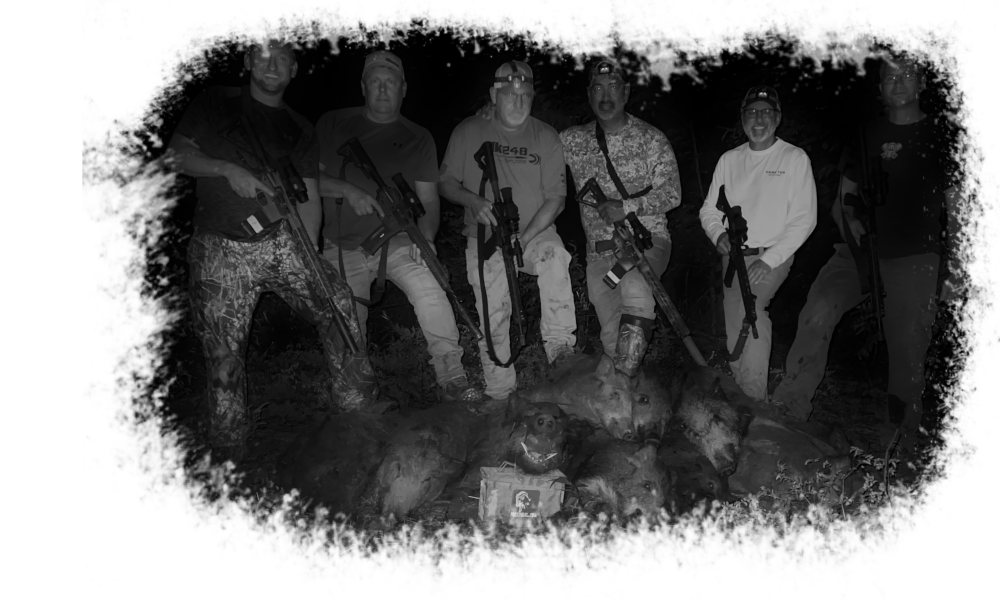 Group of six men outdoors at night, posing with five hunted wild boars and hunting rifles.