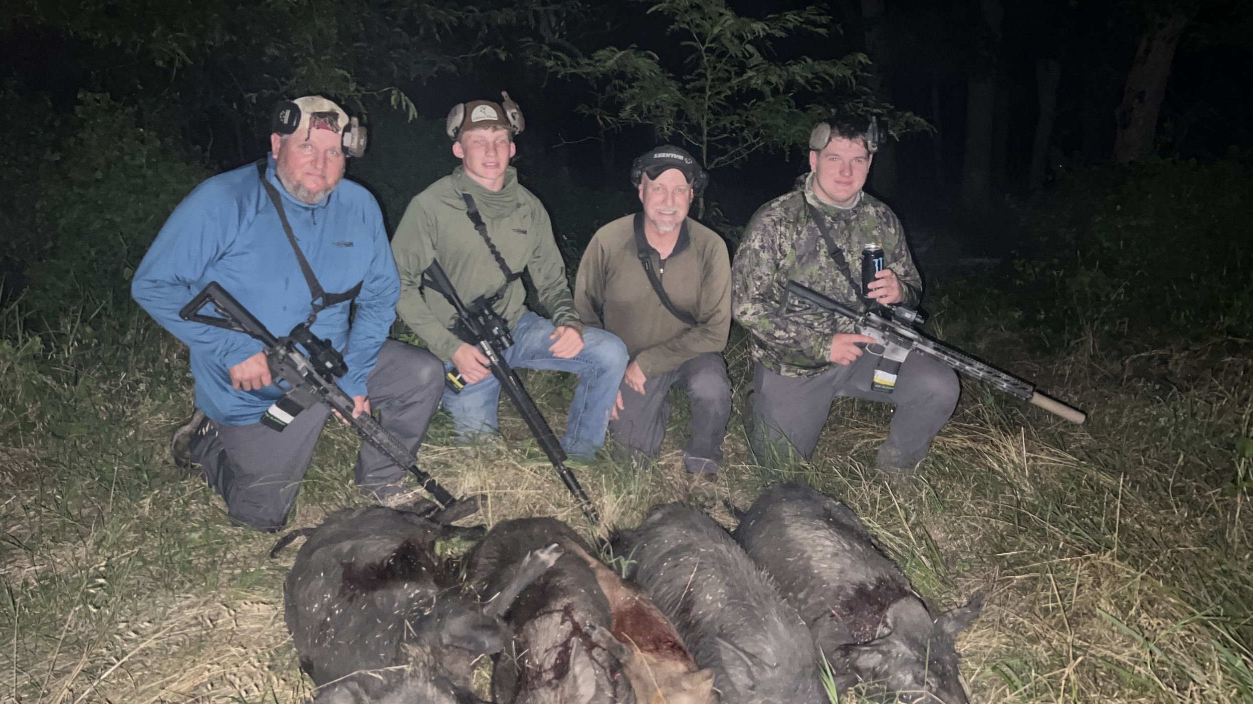 Four men kneeling outdoors at night with rifles, surrounded by logs and trees, wearing camouflage and outdoor gear.