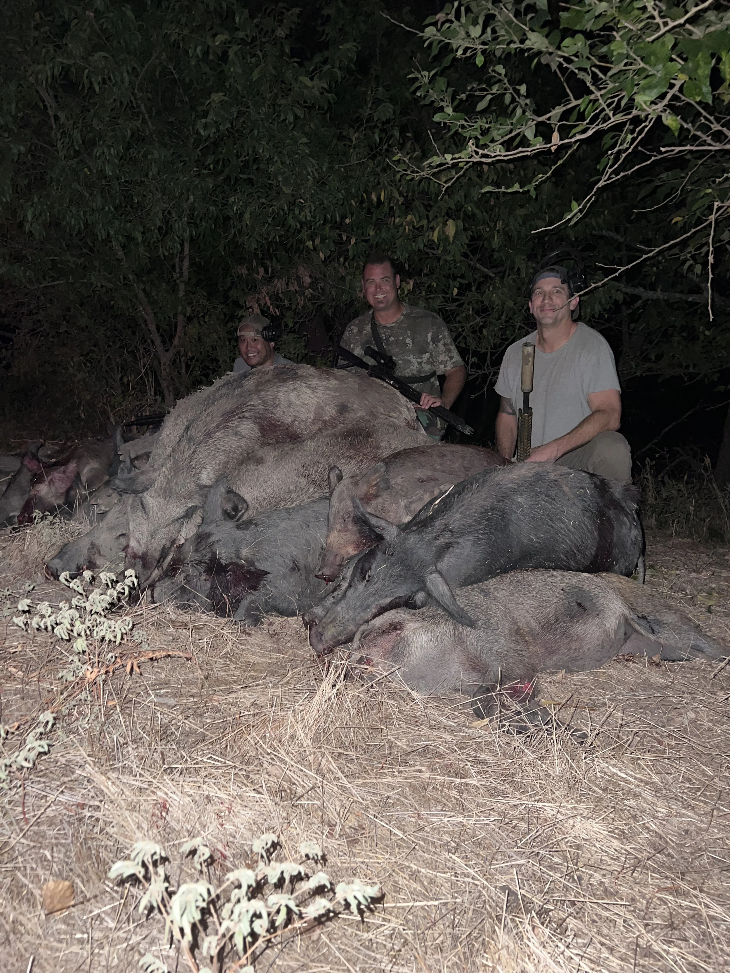 Three men in camouflage and neutral-colored clothing posing behind a large pile of pigs they have hunted, in a forest setting at night.