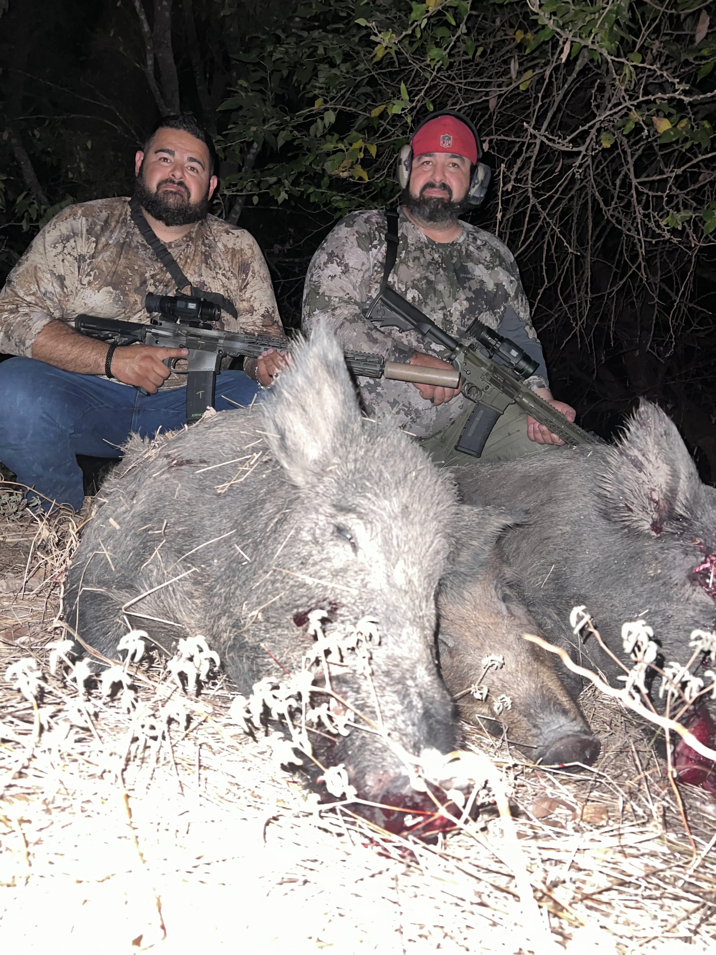 Two men sitting behind a large dead wild boar in a wooded area at night, holding rifles, with a background of trees and branches.
