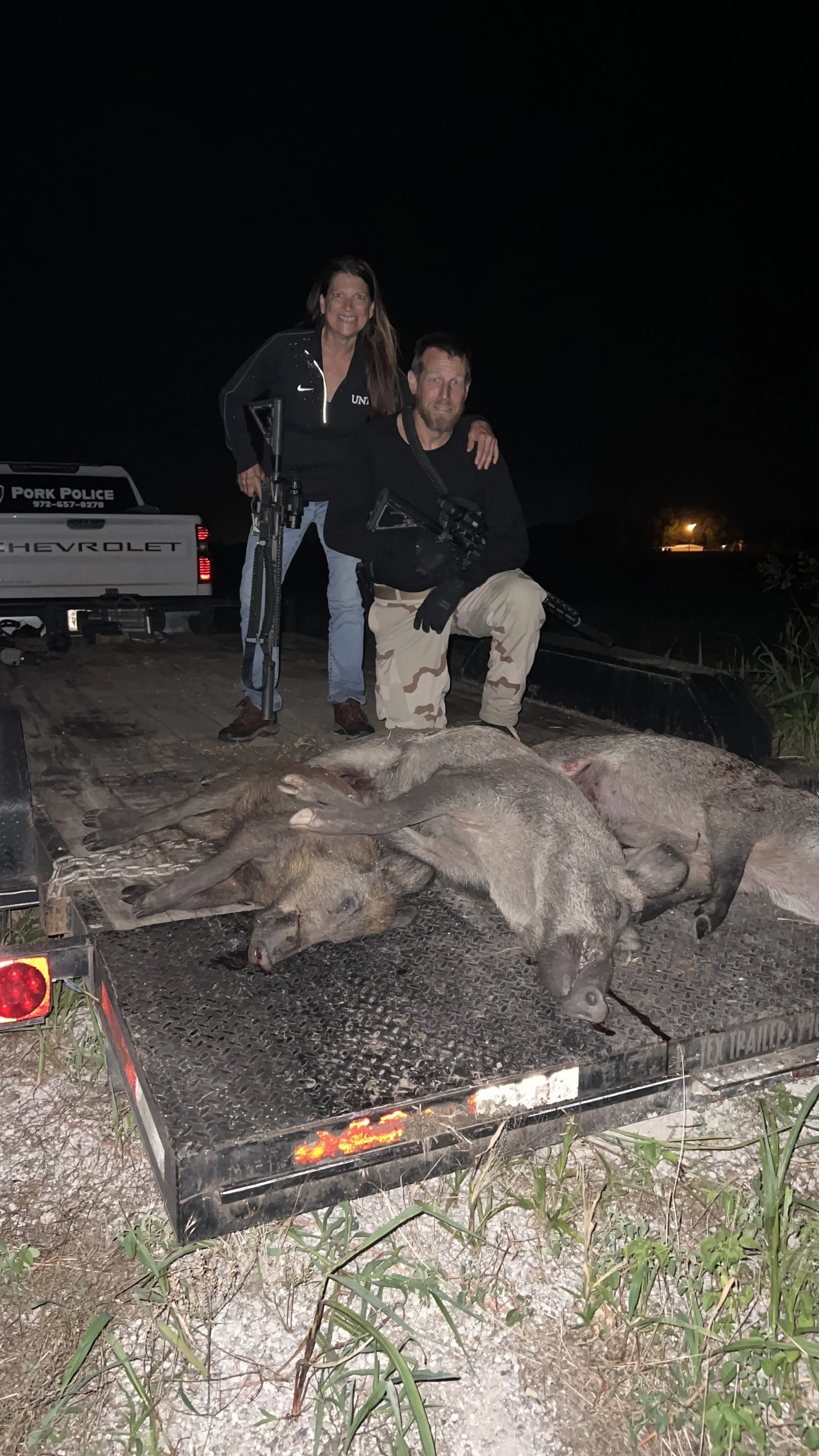A man and a woman posed with rifles on a flatbed trailer, alongside the carcasses of multiple wild pigs, in a nighttime outdoor setting.