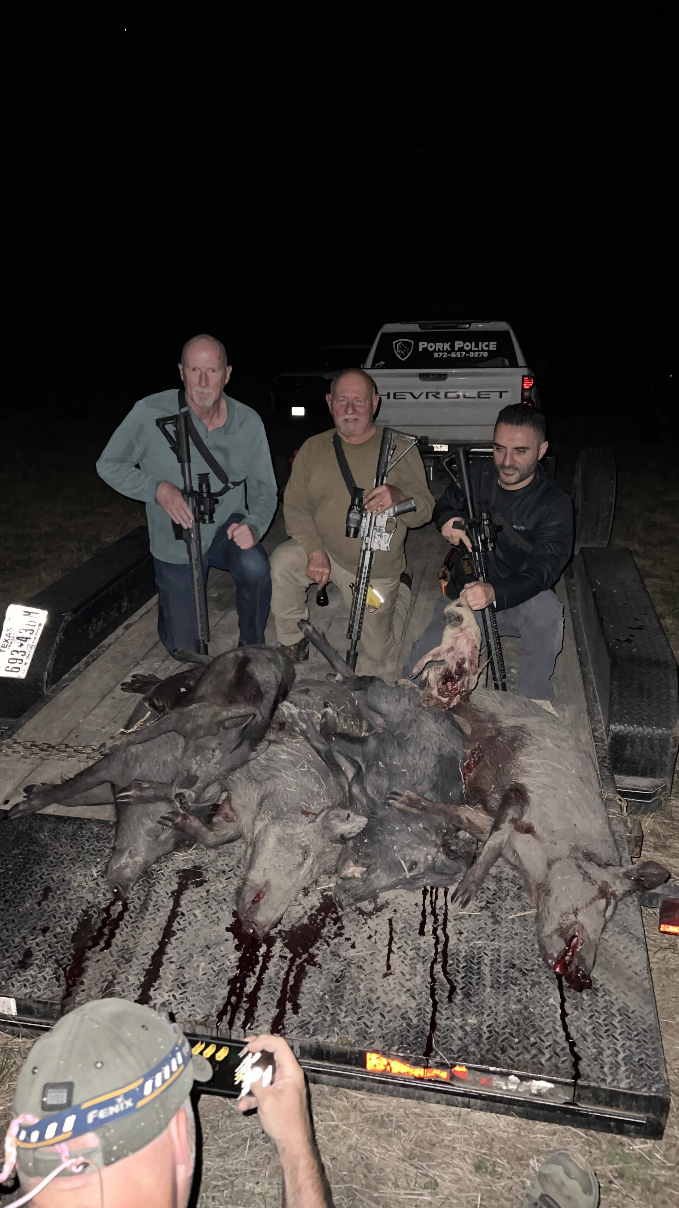 Three men on a trailer with multiple dead hogs, holding rifles, after a night of hunting at night outside with a pickup truck in the background, labeled 'Pork Police'.