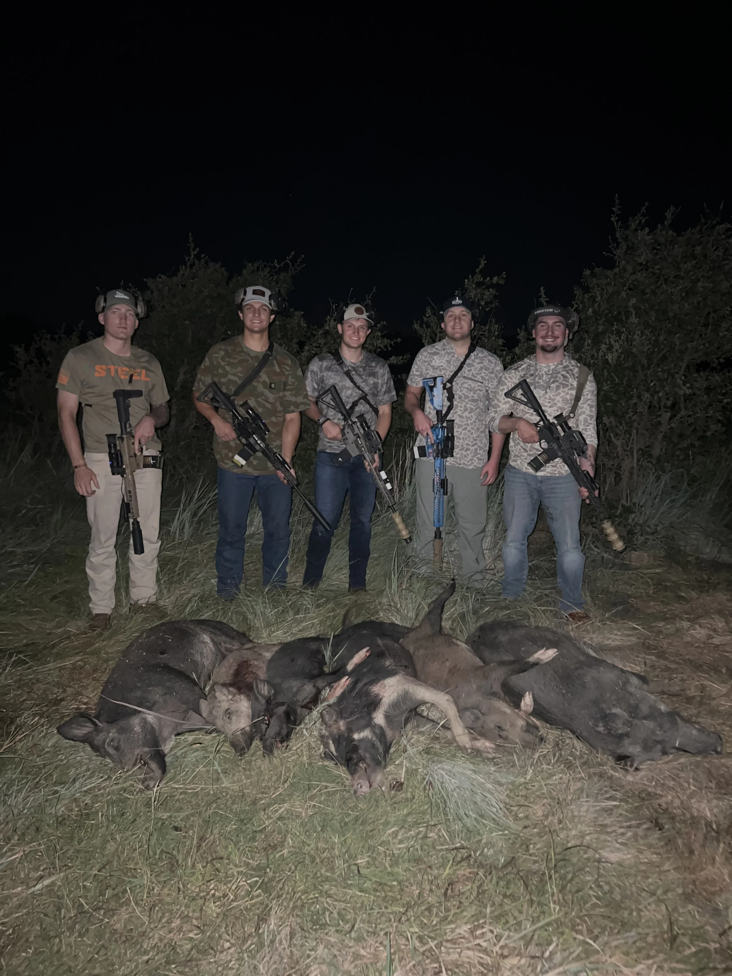 Five people standing outdoors at night with rifles, posing behind a line of pig carcasses on the ground.