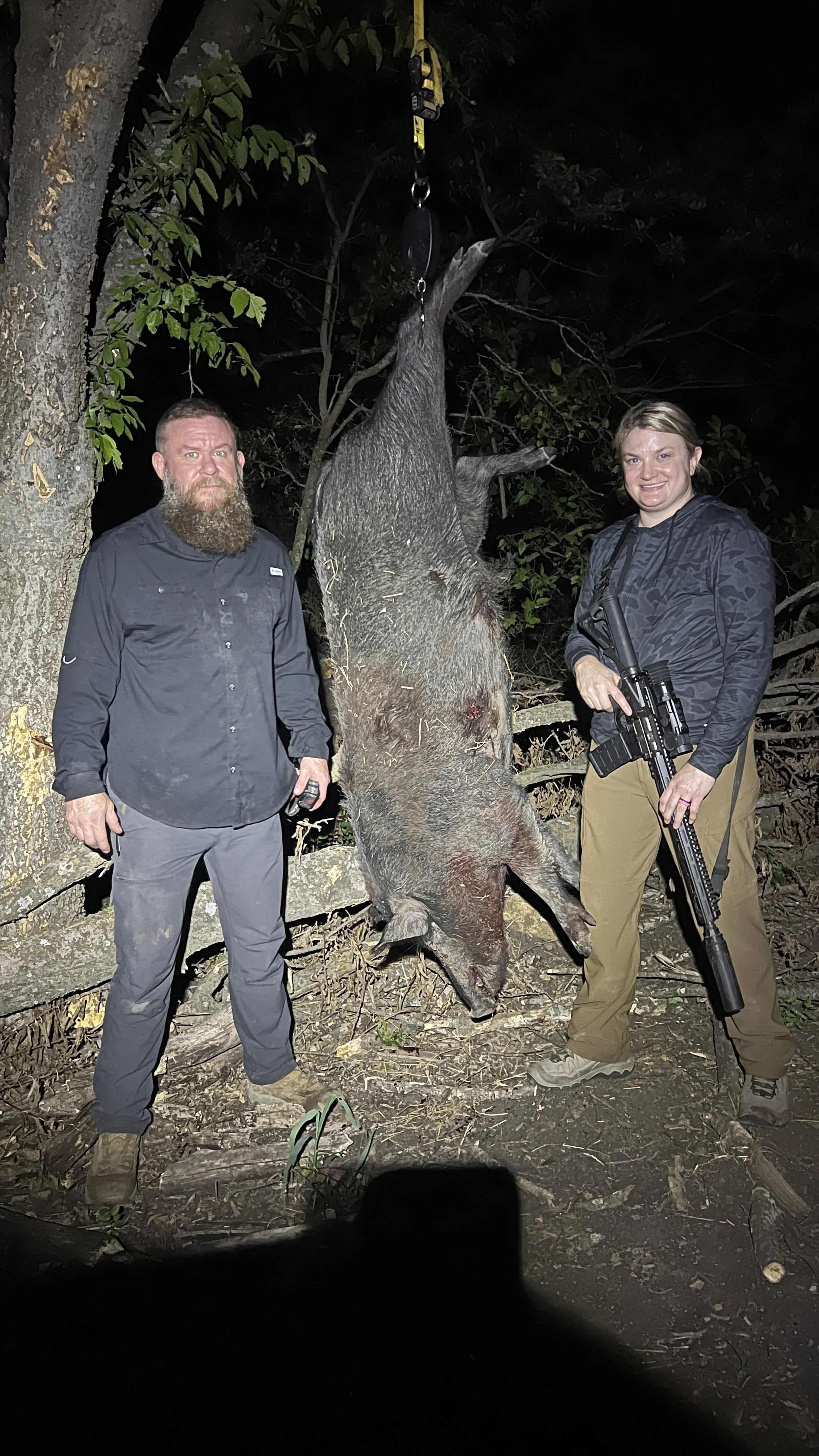 Two hunters stand on either side of a large, freshly killed boar hanging upside down from a tree. The hunter on the right is holding a rifle, and the hunter on the left is holding a device, both posing for the photo at night.