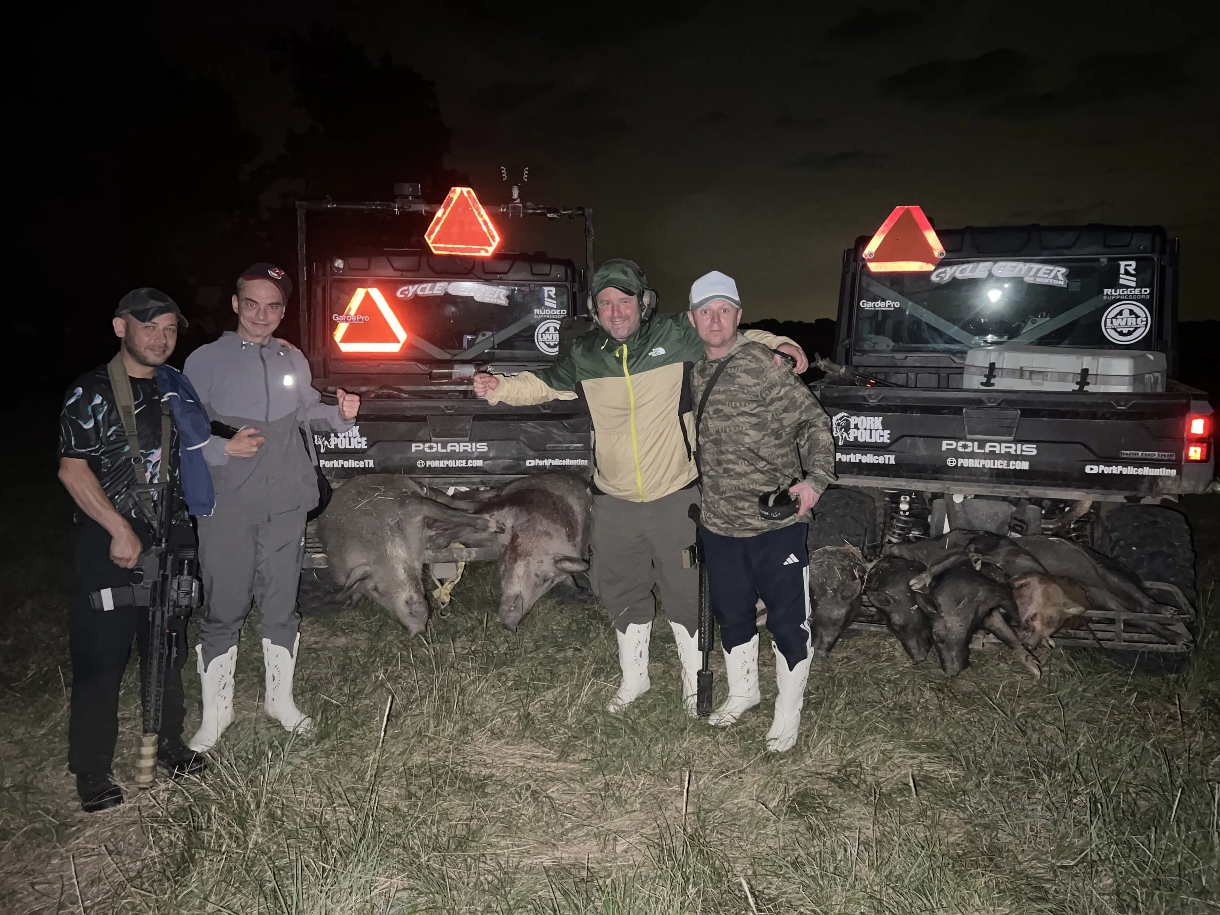 Four men standing in front of utility vehicles at night; two are holding deer carcasses, wearing white snow boots, camo and outdoor gear. The vehicles have reflective safety triangles and stickers indicating police and hunting logos.