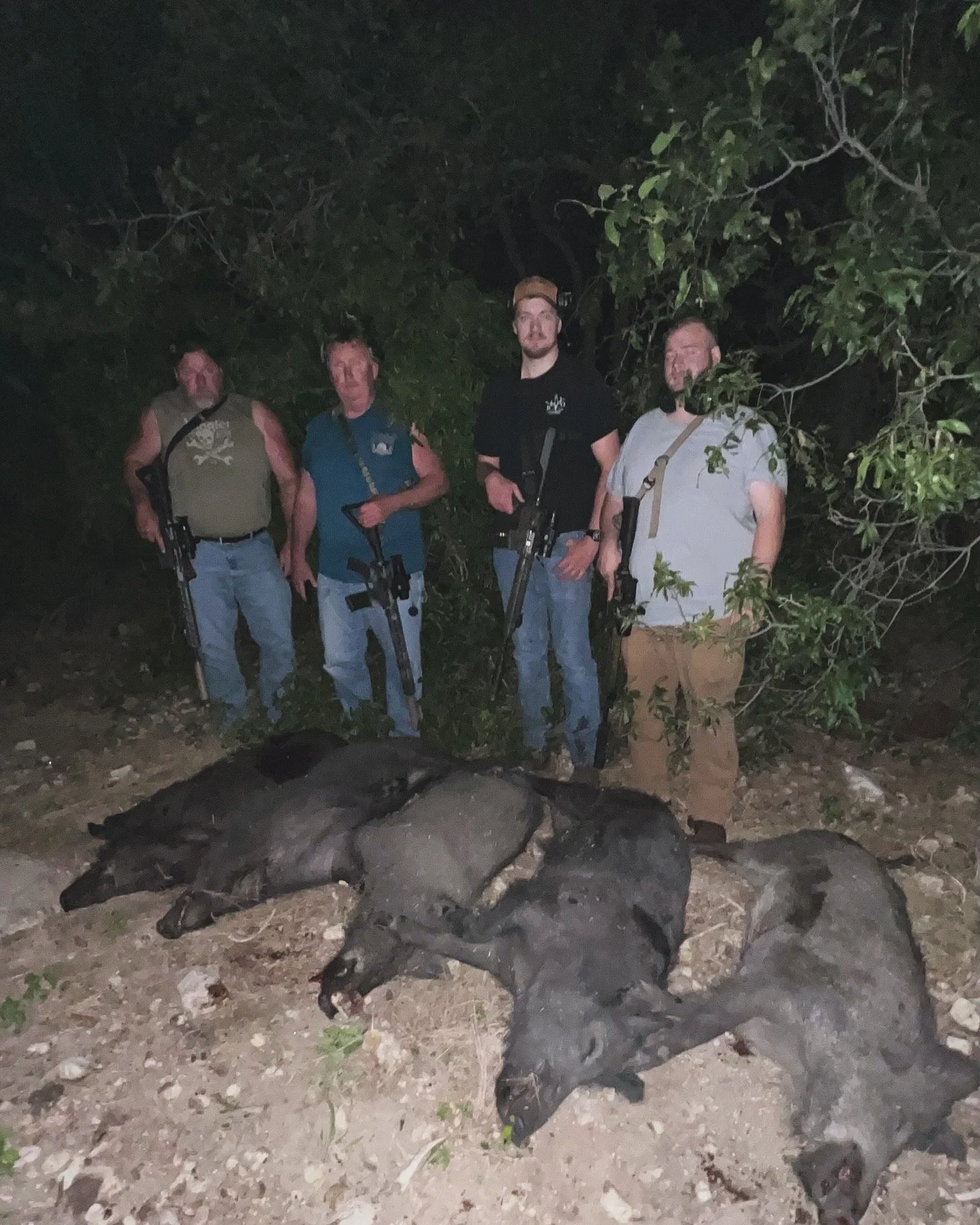 Four men standing outdoors at night, dressed in casual clothes, holding rifles, and posing beside a large black dead animal on the ground, with trees and rocks in the background.