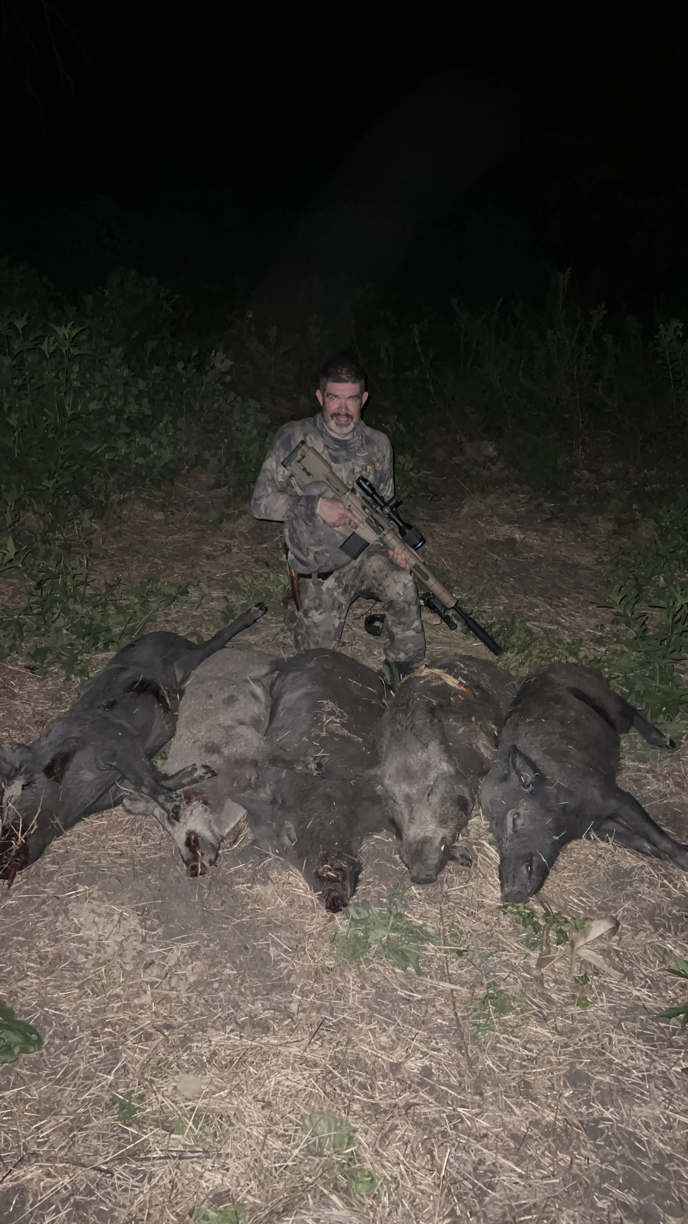 A man in camouflage clothing kneeling outdoors at night, holding a scoped rifle, with several dead pigs laid out in front of him on the ground.