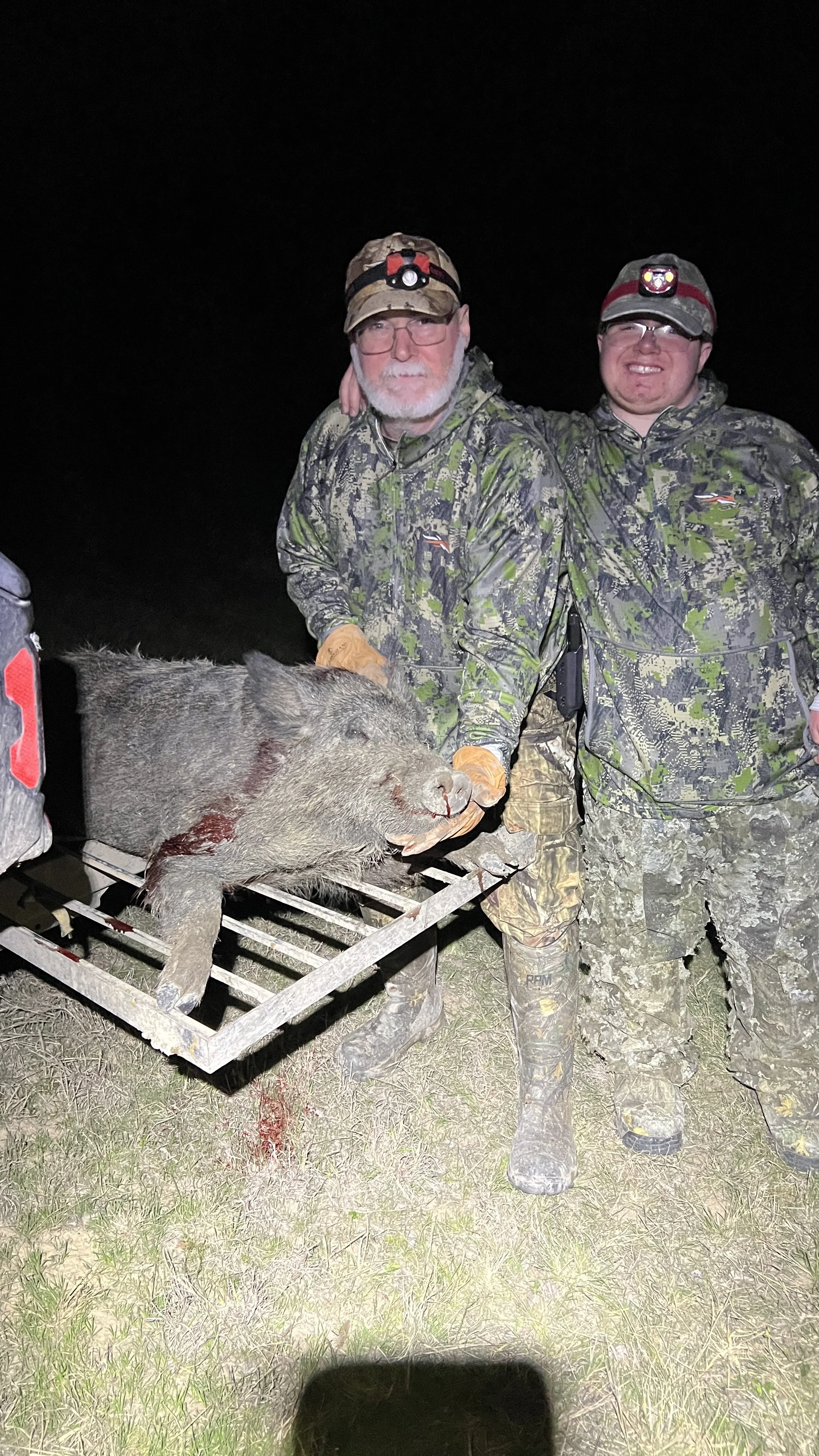 Two men in camouflage clothing and hats standing at night. They are holding a large dead wild boar on a metal grate, some blood visible on the animal. One man has his arm around the other's shoulder.