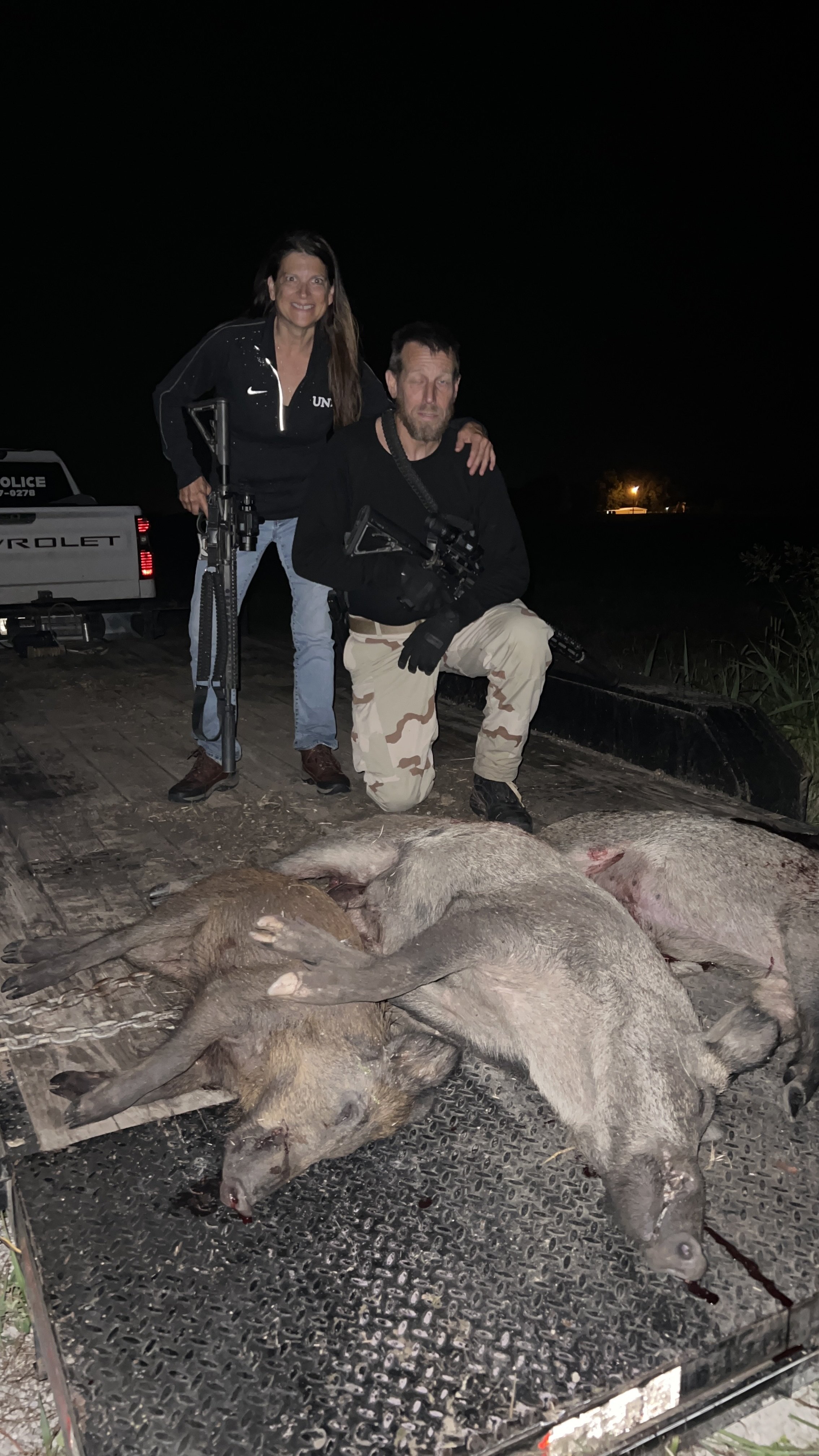 Two individuals, one woman and one man, are kneeling and standing behind a group of dead pigs on a truck bed at night. The woman is smiling and wearing a black jacket with a Nike logo, and the man is holding a rifle, wearing a black shirt and camoufl