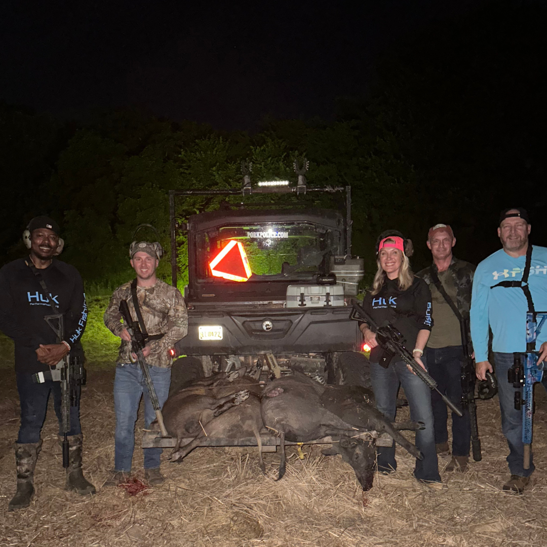 Six people standing behind a group of dead pigs in front of an off-road vehicle at night.