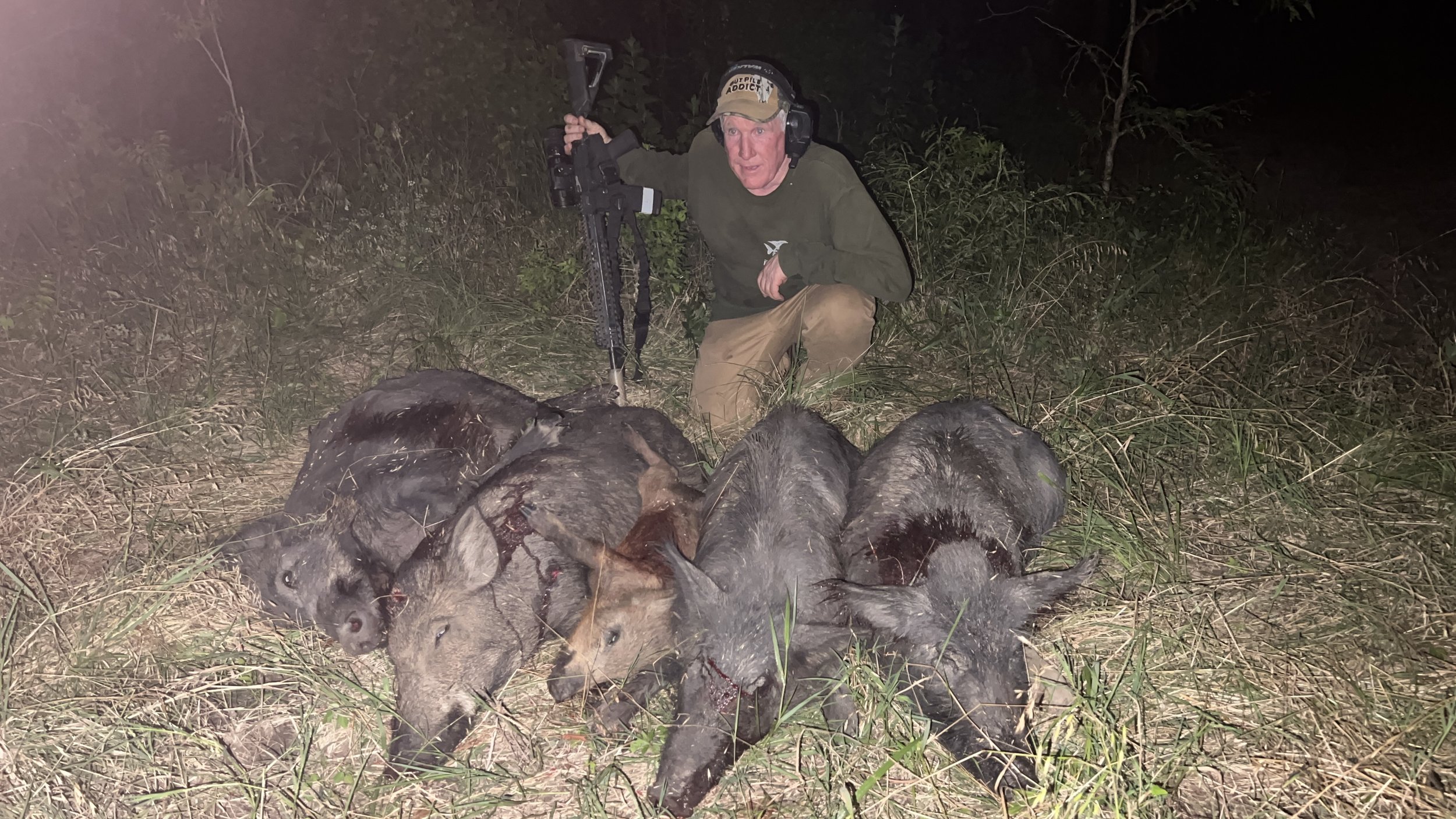 A man kneeling in a grassy outdoor area at night with a hunting rifle, surrounded by multiple dead wild hogs.