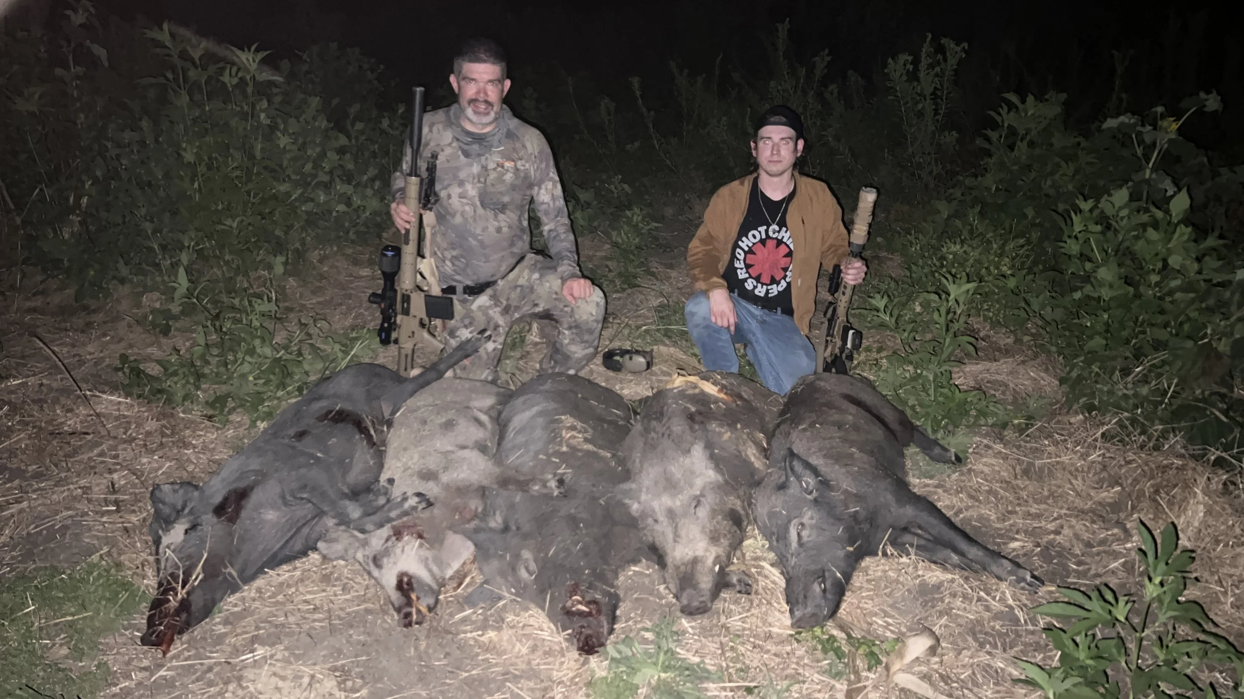 Two men kneeling behind a pile of dead animals, possibly wild boar or pig, in a forested area at night, holding rifles and posing for the photo.