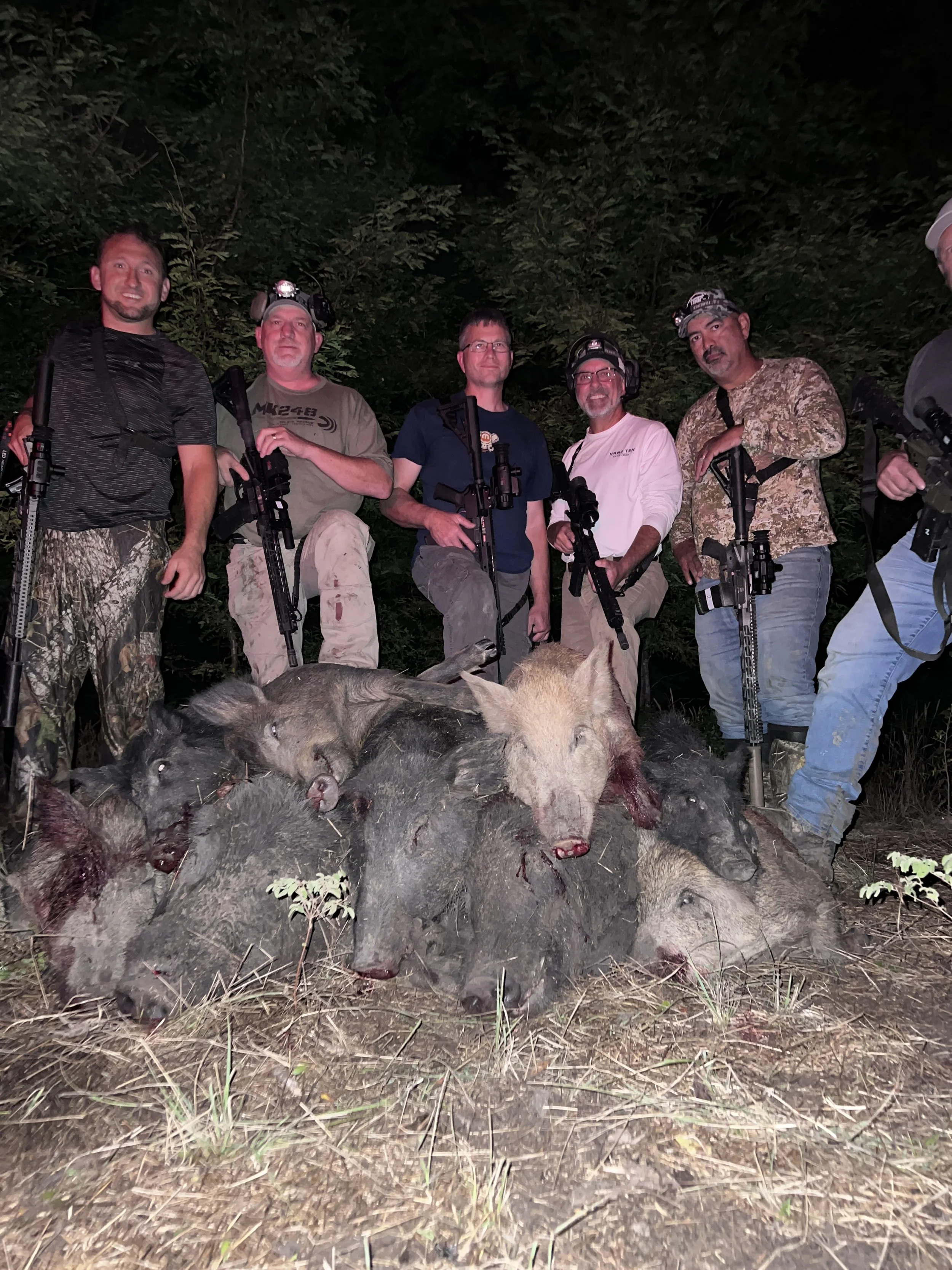 A group of six people standing outdoors at night in front of trees, holding rifles, with a pile of dead boars in front of them on the ground.