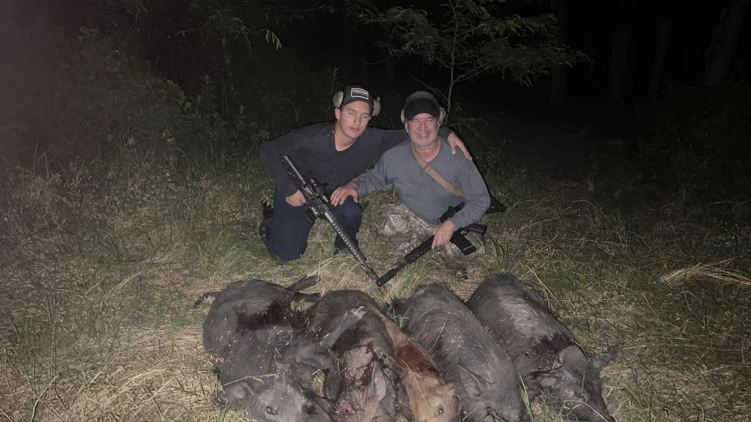 Two men wearing protective earmuffs and dark clothing sitting on the ground outdoors at night, with a pile of logs in the foreground and trees in the background.