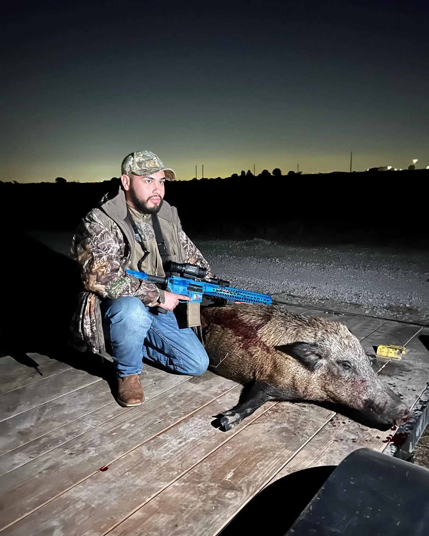 A man in camouflage clothing kneels next to a large pig lying on a wooden platform, with a hunting rifle resting on the pig's side, outdoors at night under a dark sky.