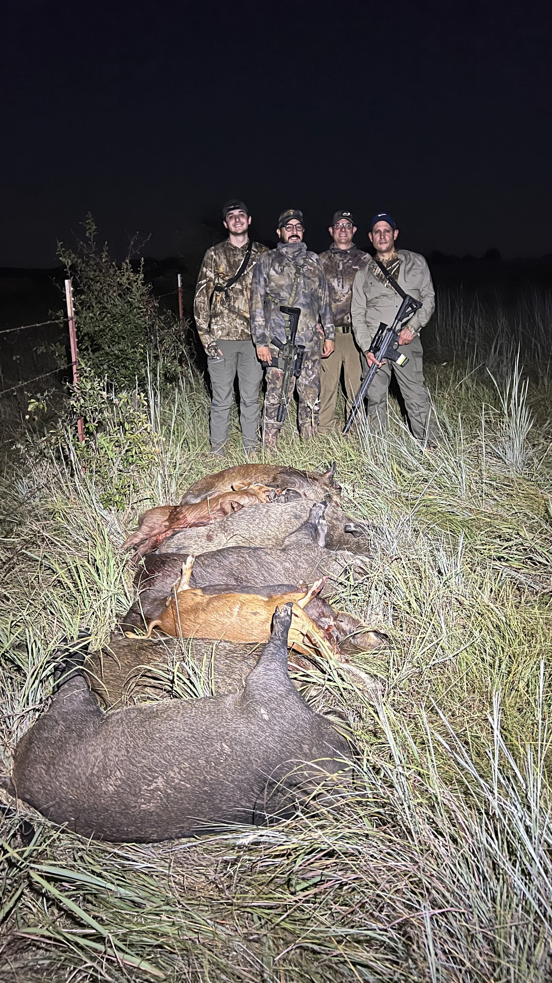 Four hunters wearing camouflage and outdoor clothing stand behind a line of dead hogs lying in tall grass at night, holding rifles.