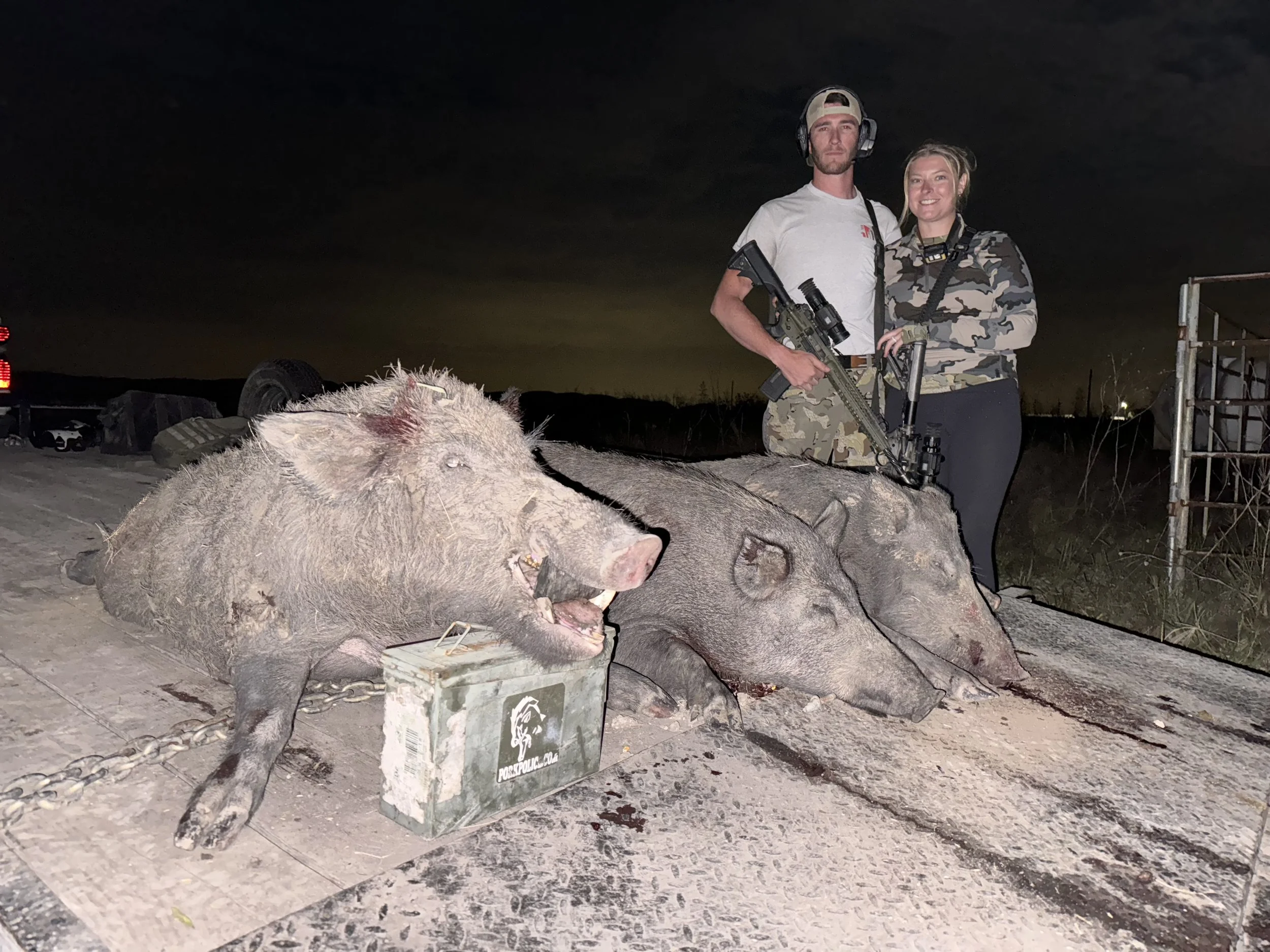 Two people standing outdoors at night, one man and one woman, both dressed in camouflage clothing and holding rifles. There are dead pigs lying on the ground in front of them, and a sign with a wildlife agency logo nearby.