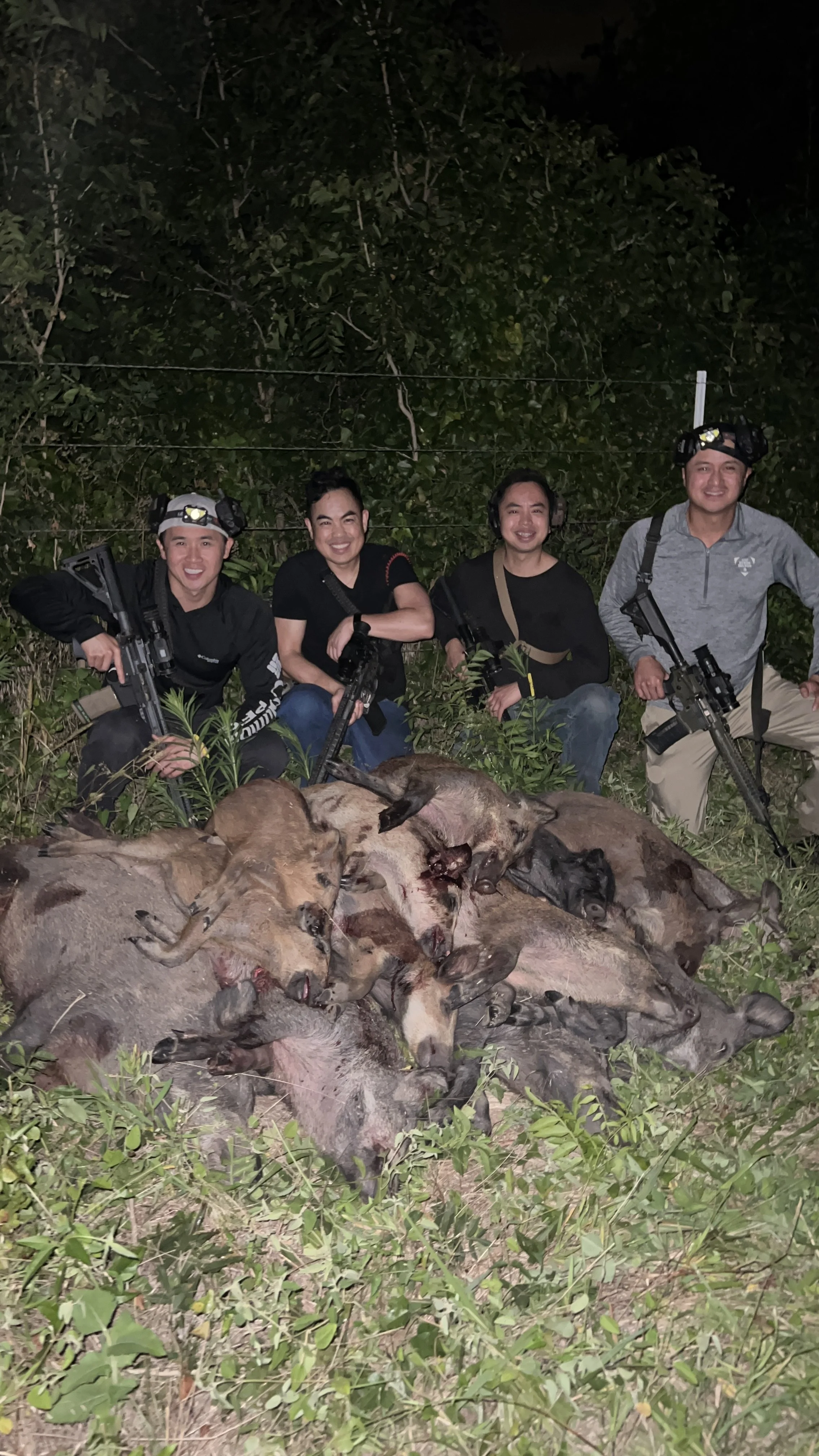 Group of four hunters with rifles kneeling behind a large pile of dead animals, mainly wild boars, in a wooded area at night.