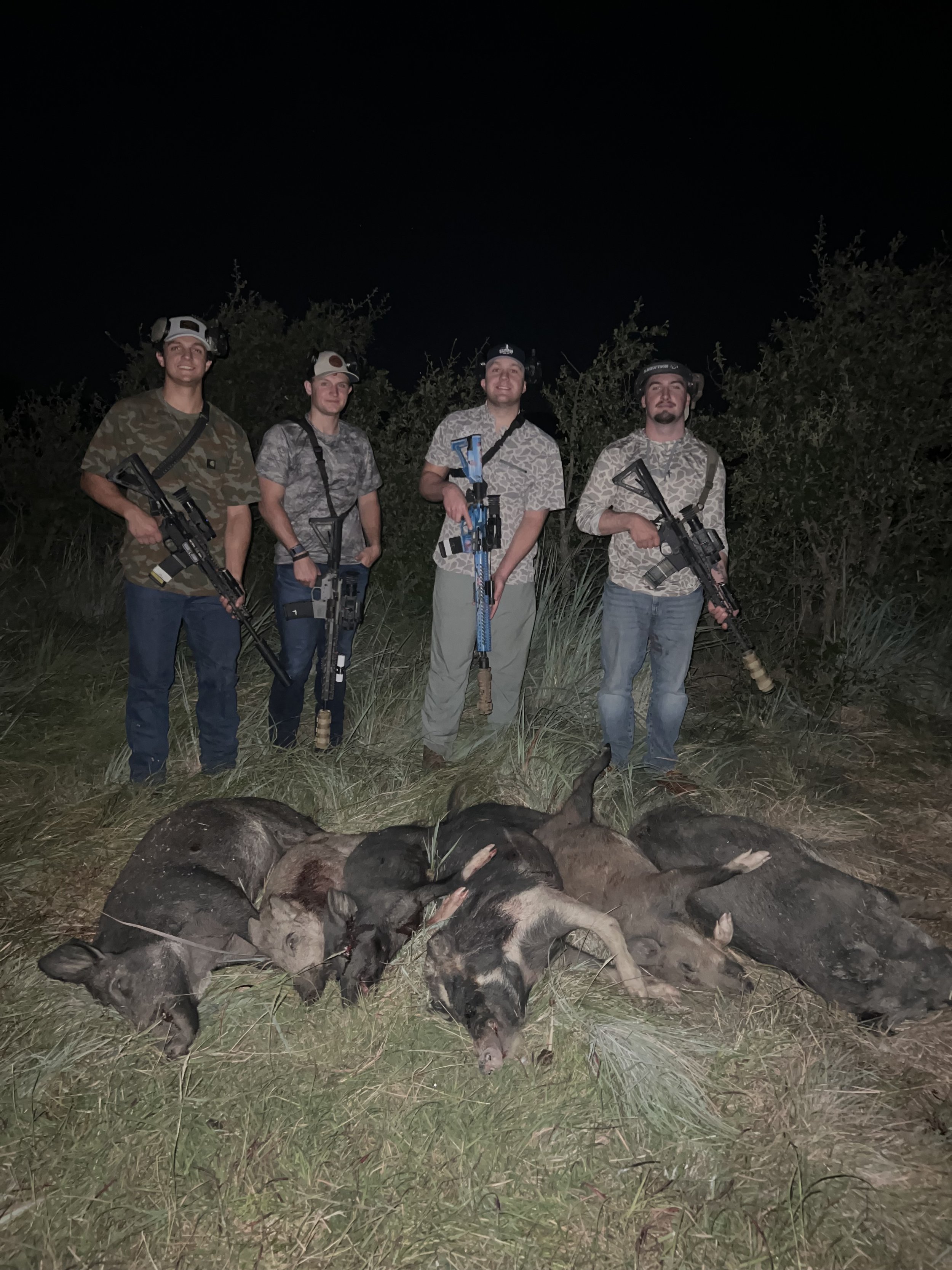 Four people standing outdoors at night, dressed in camouflage shirts and holding rifles, posing behind a line of dead wild boars lying on the ground.