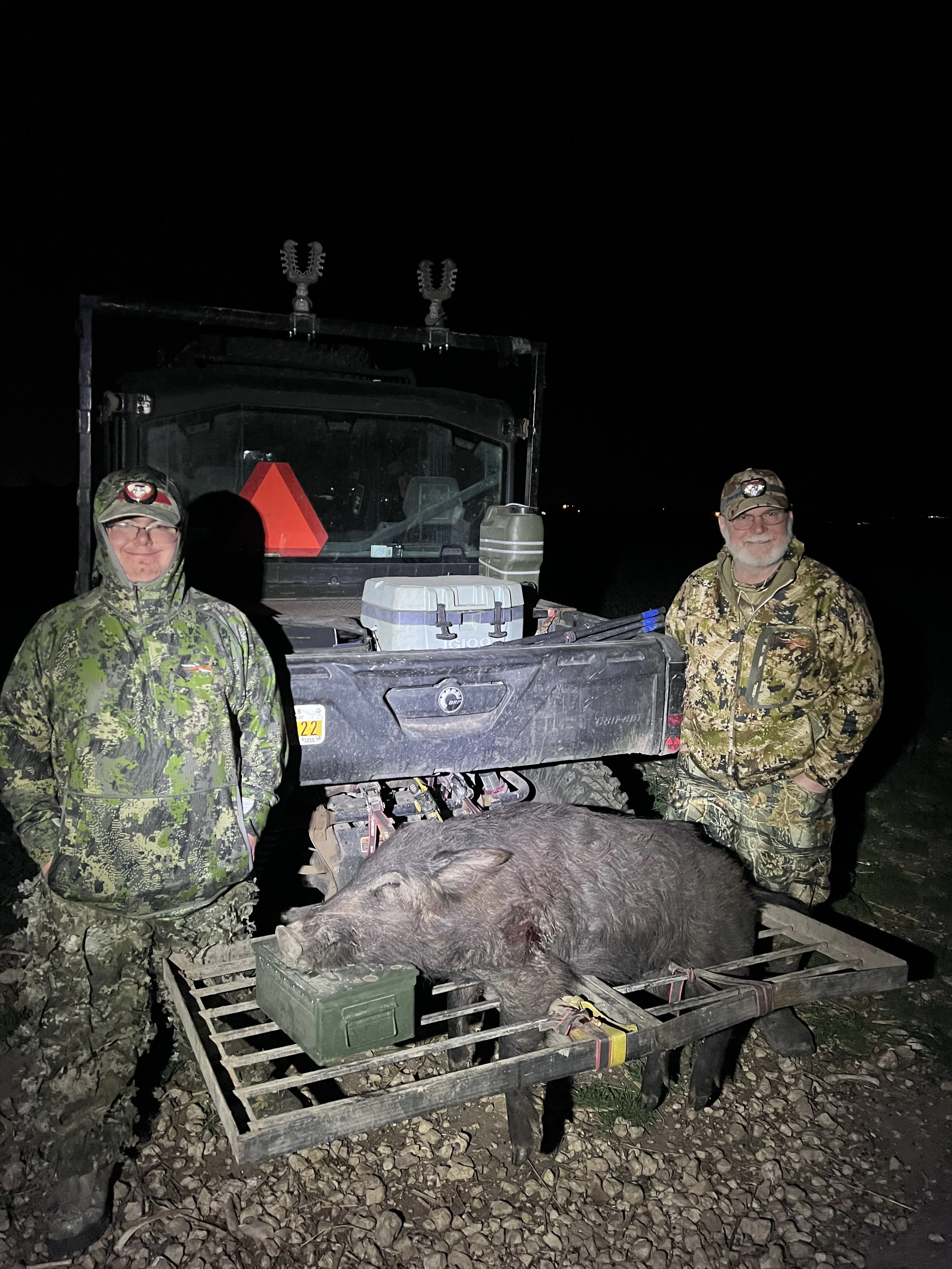 Two men in camouflage clothing stand on either side of a large wild hog lying on a metal cart, with a utility vehicle in the background at night.