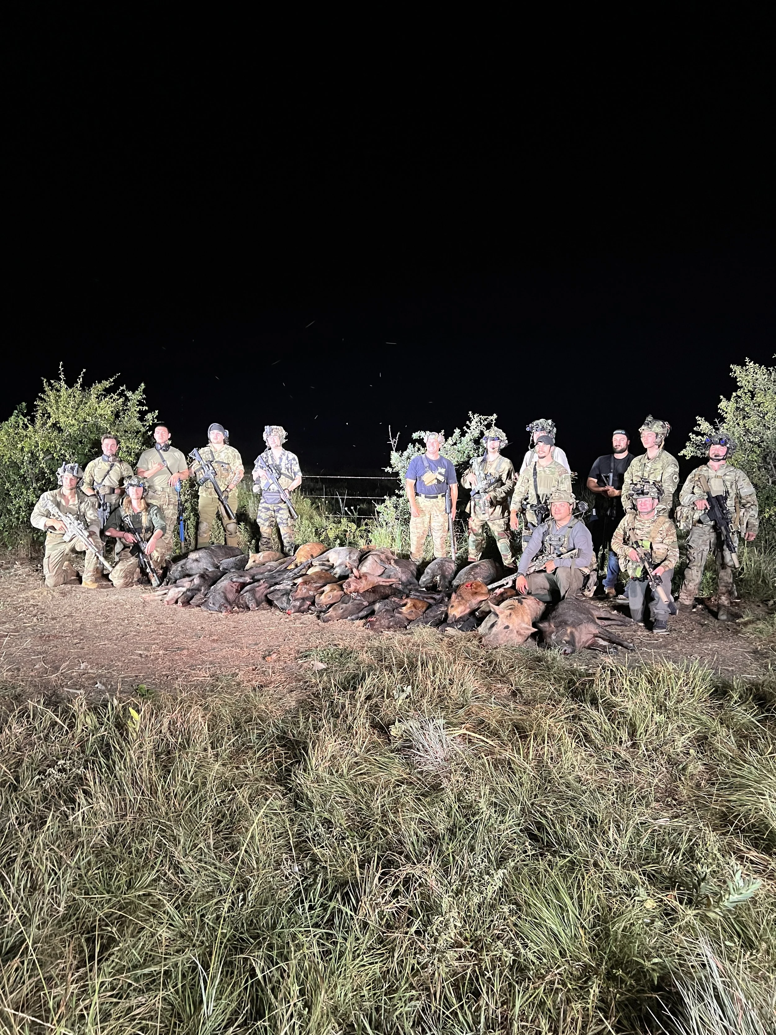 A group of soldiers in military uniforms and gear standing together outdoors at night, posing with dead animals laid on the ground in front of them.