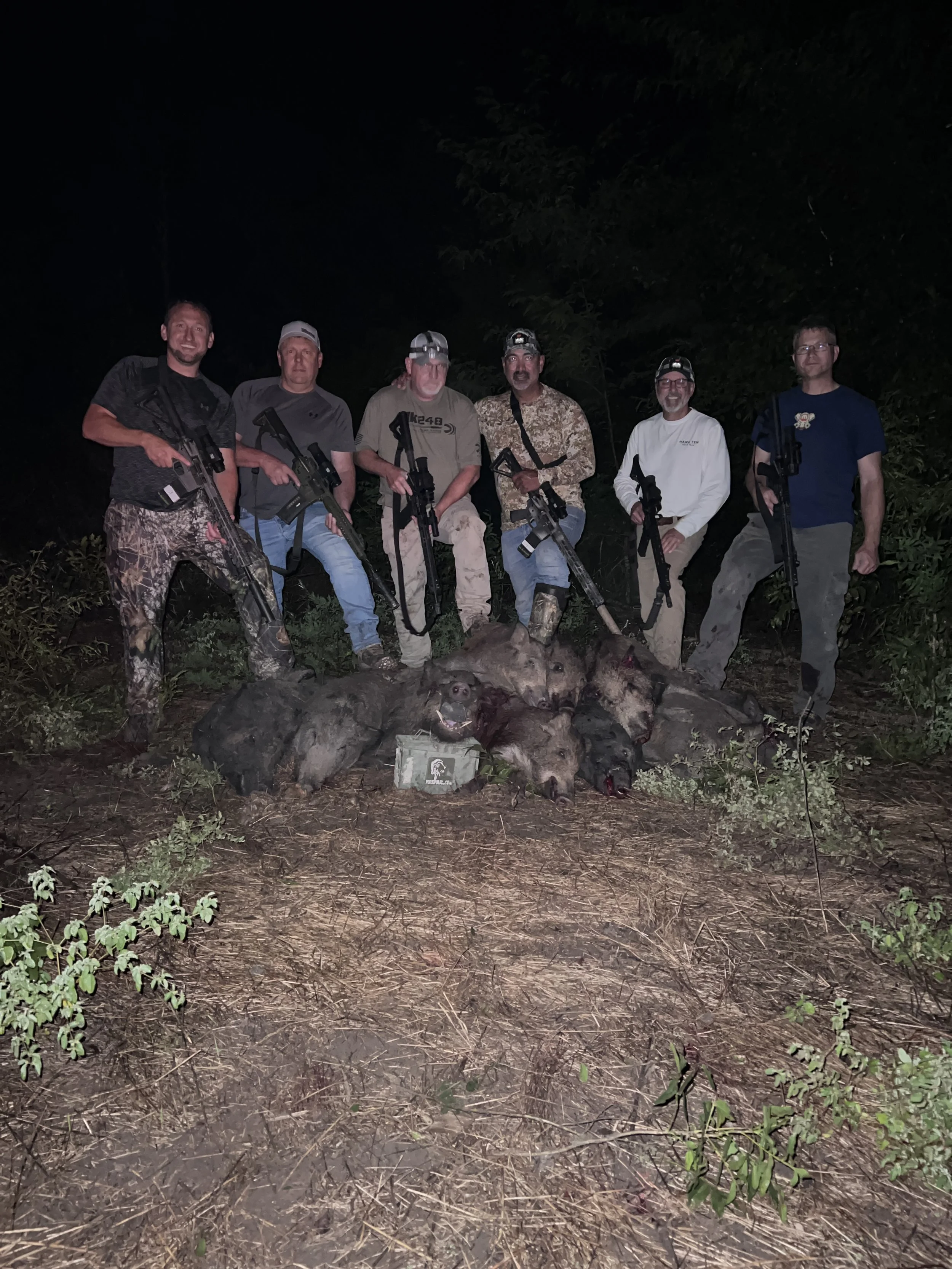 Group of six people at night outdoors, posing with rifles, with dead wild boar on the ground in front of them.