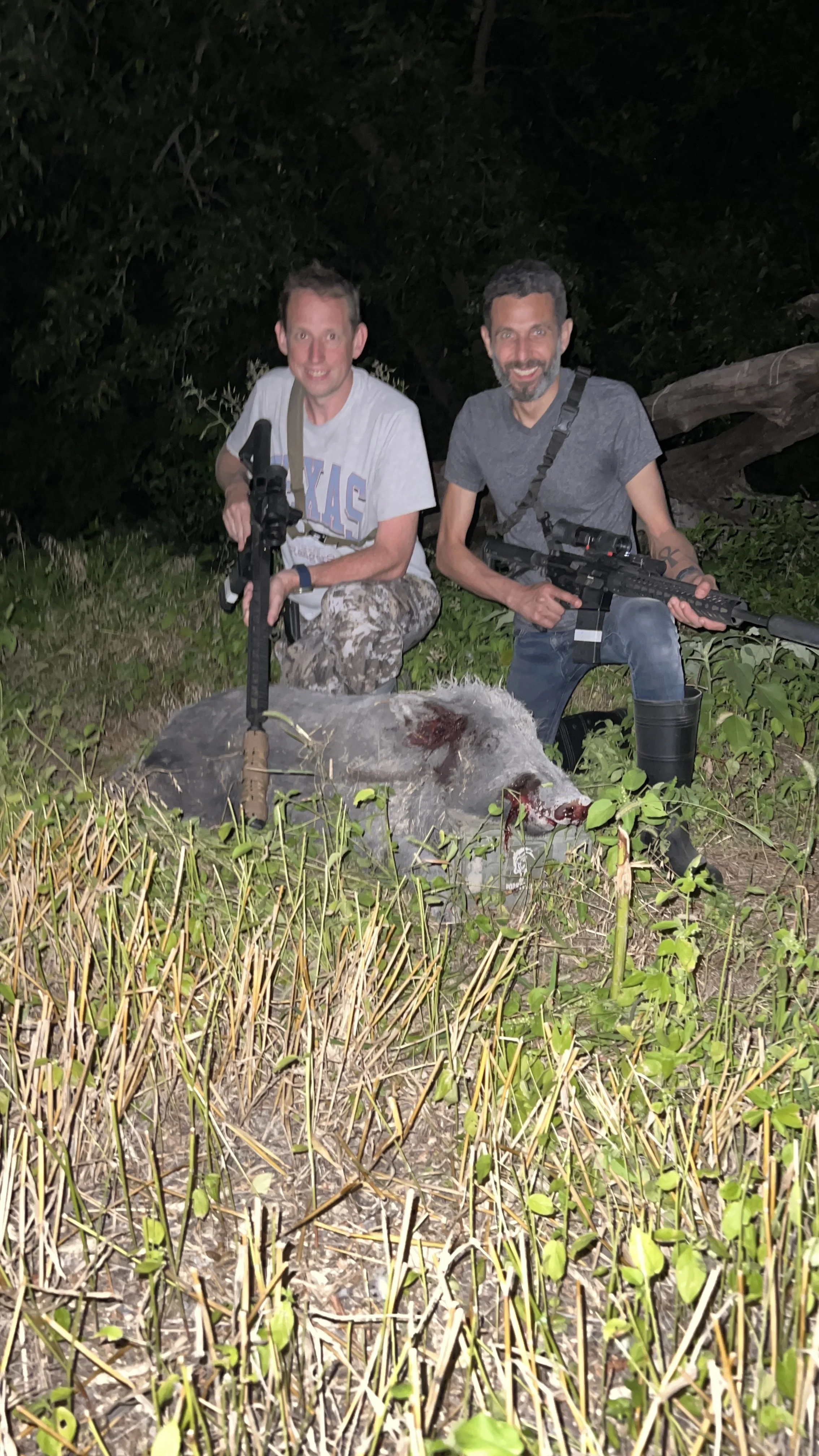 Two men kneeling beside a dead boar in a grassy area at night, holding rifles and smiling at the camera.