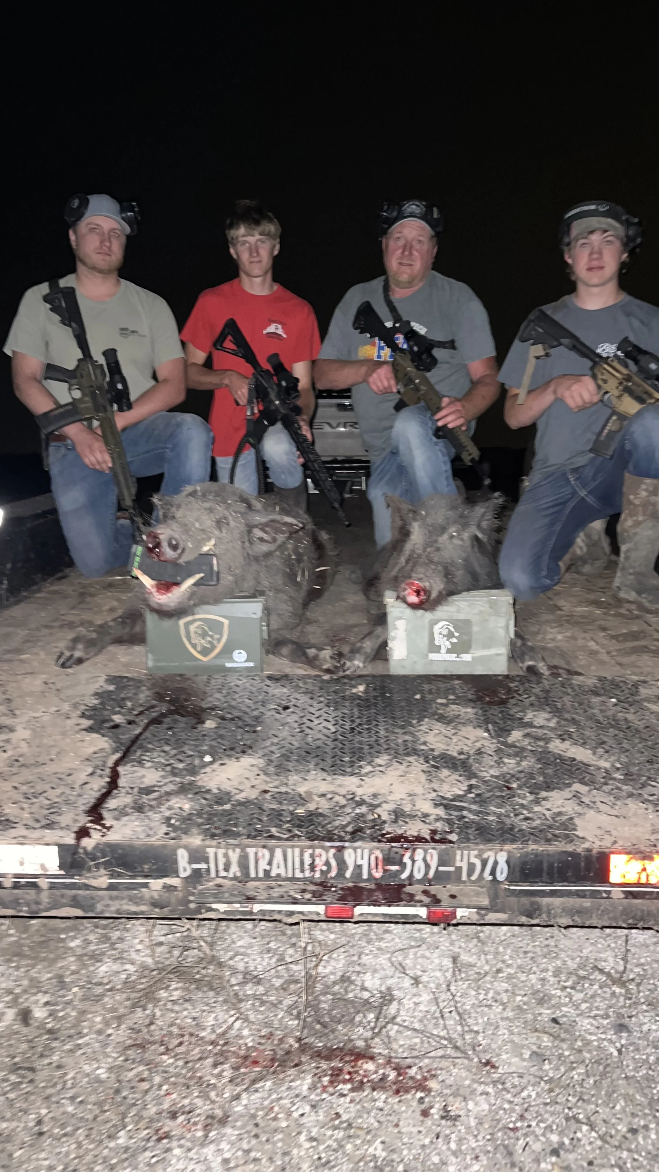 Four men wearing tactical gear and holding rifles kneeling on a flatbed trailer with two deceased boars in front of them at night. The trailer has a contact number and the company name 'B- Tex Trailers.'