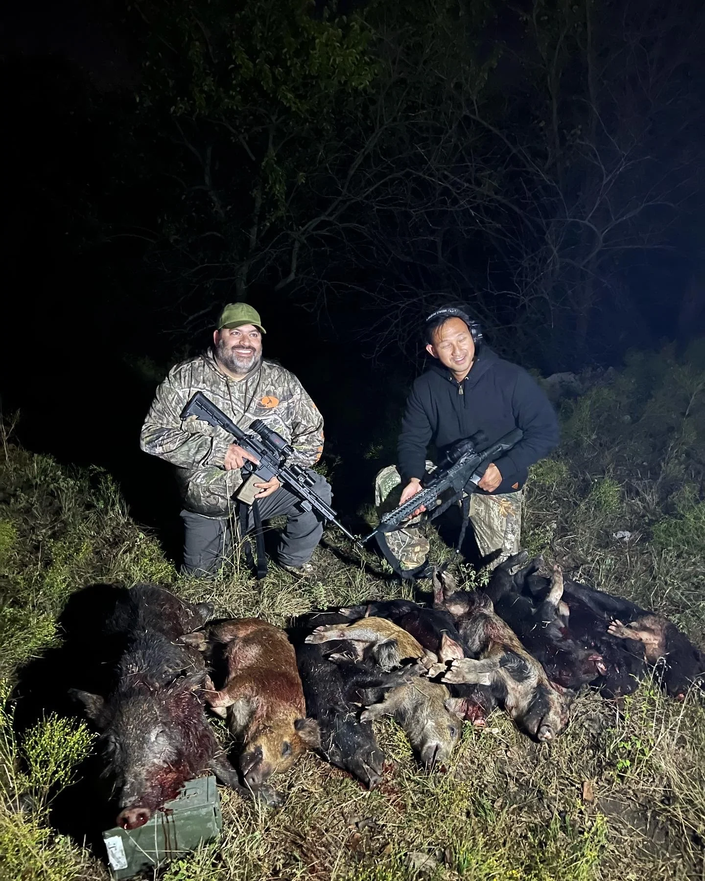 Two men smiling and posing with rifles next to a large pile of hunted animals at night outdoors, with trees and grass in the background.