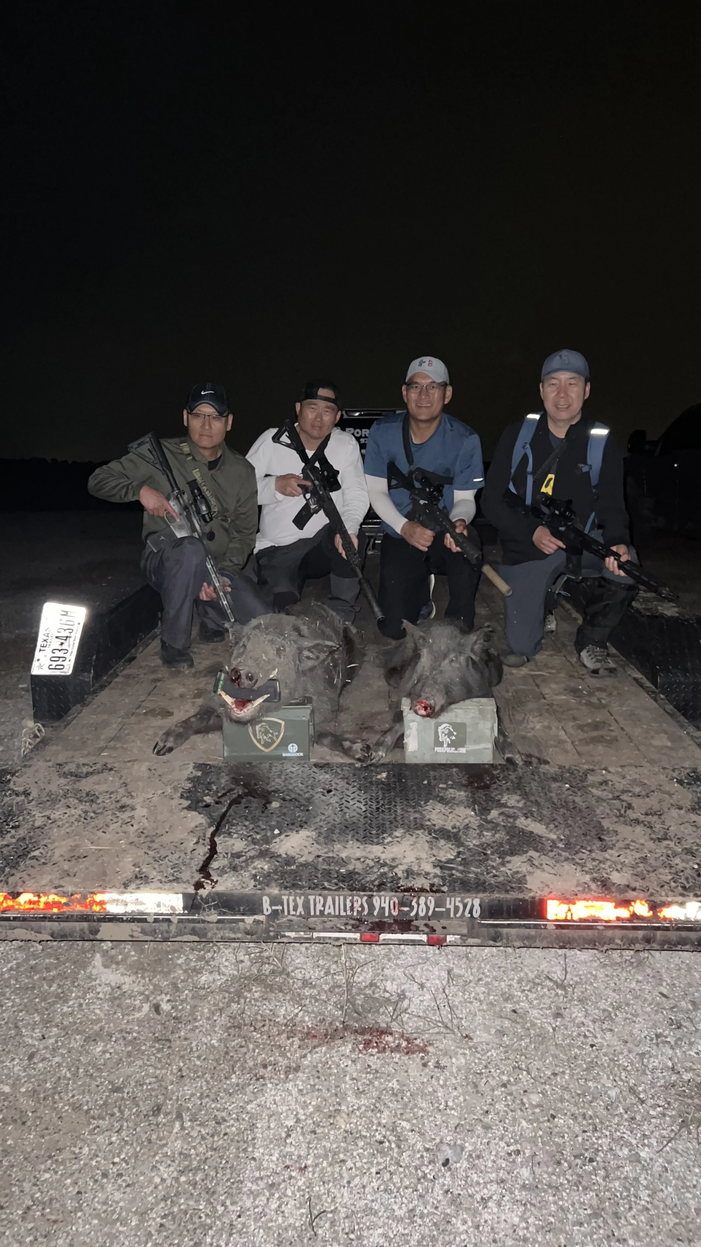 Four men kneeling on a trailer holding rifles, with two dead hogs in front of them. The photo was taken at night, and there is a Texas license plate visible on the trailer.
