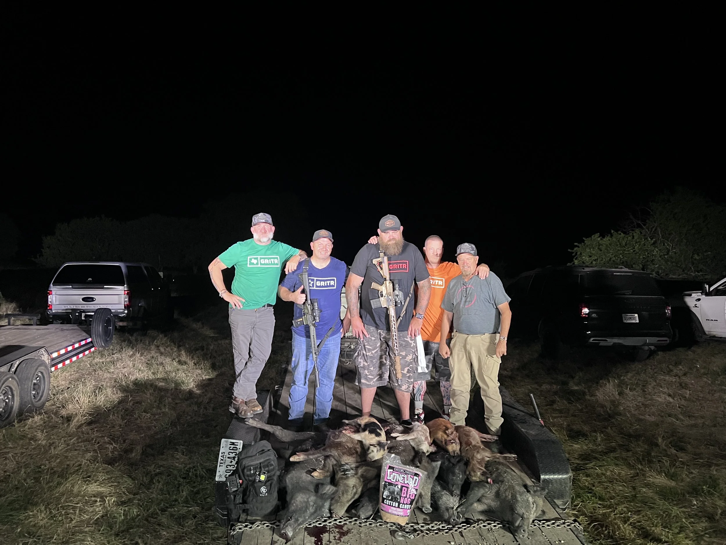 Group of five men standing outdoors at night, posing with their arms around each other. In front of them on a table are multiple hunted game animals, dog tags, and a magazine. The background shows parked vehicles and trees under a dark sky.