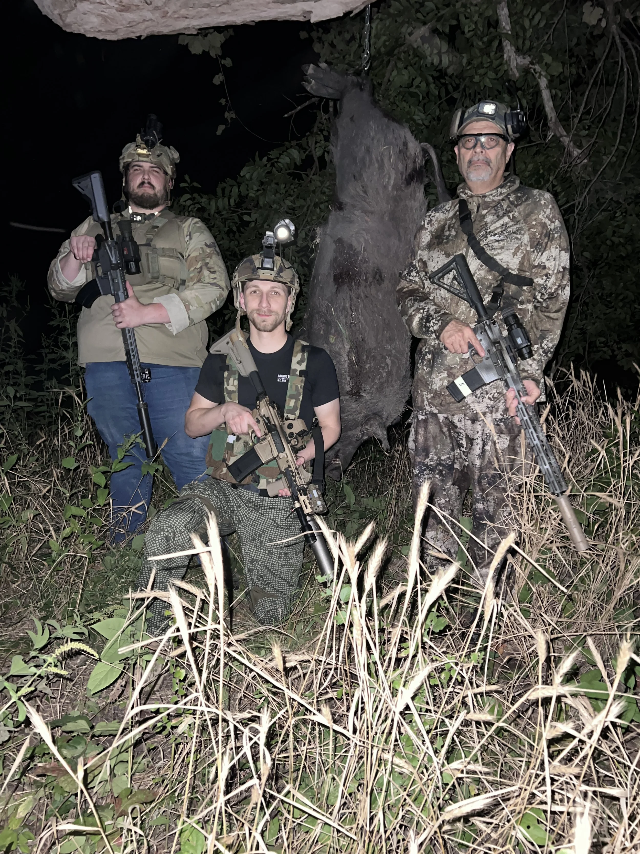 Three men in camouflage and tactical gear, two holding rifles, standing in a dense, dark outdoor area with tall grass and trees, with a large animal hanging from a tree.