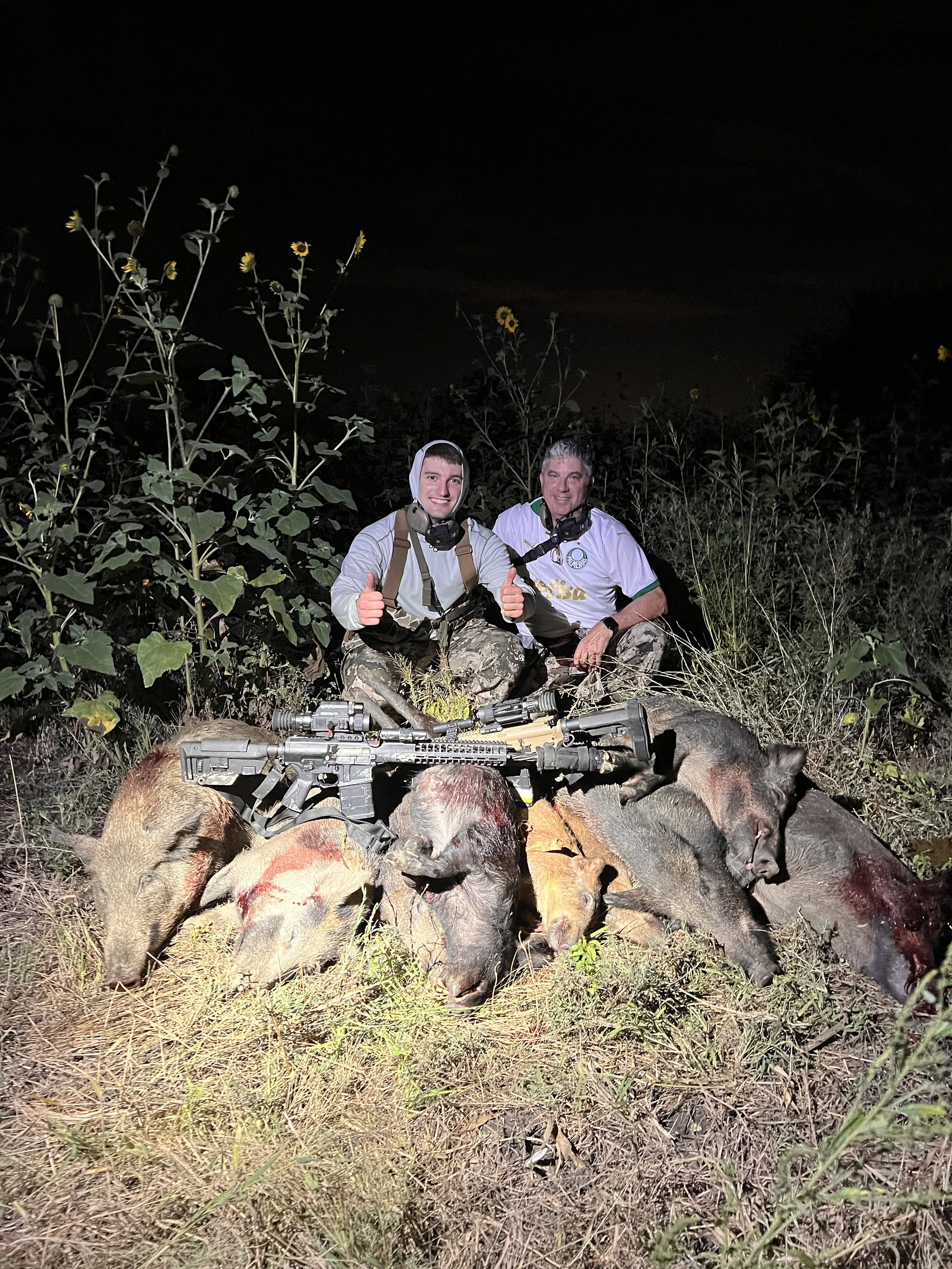 Two hunters sitting in a field at night behind a pile of hunted wild boar carcasses with rifles laid on top, surrounded by tall plants and sunflowers, smiling and giving thumbs-up.