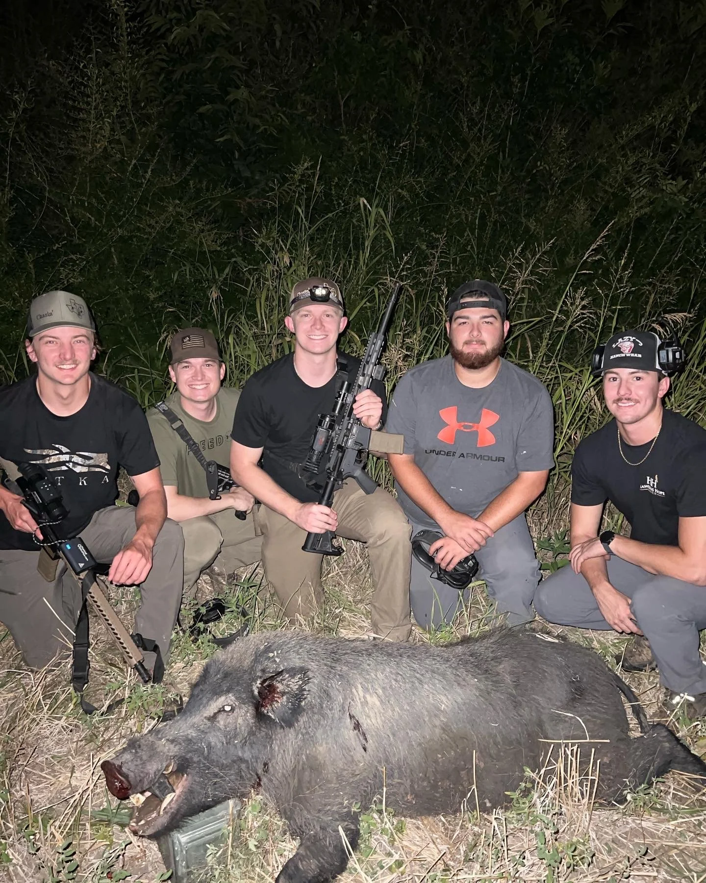 Five young men kneeling and sitting in a grassy area at night, posing with a recently hunted wild boar. They are dressed in casual outdoor clothing, some with hats and headlamps, and one is holding a rifle. The boar is large with visible wounds and i