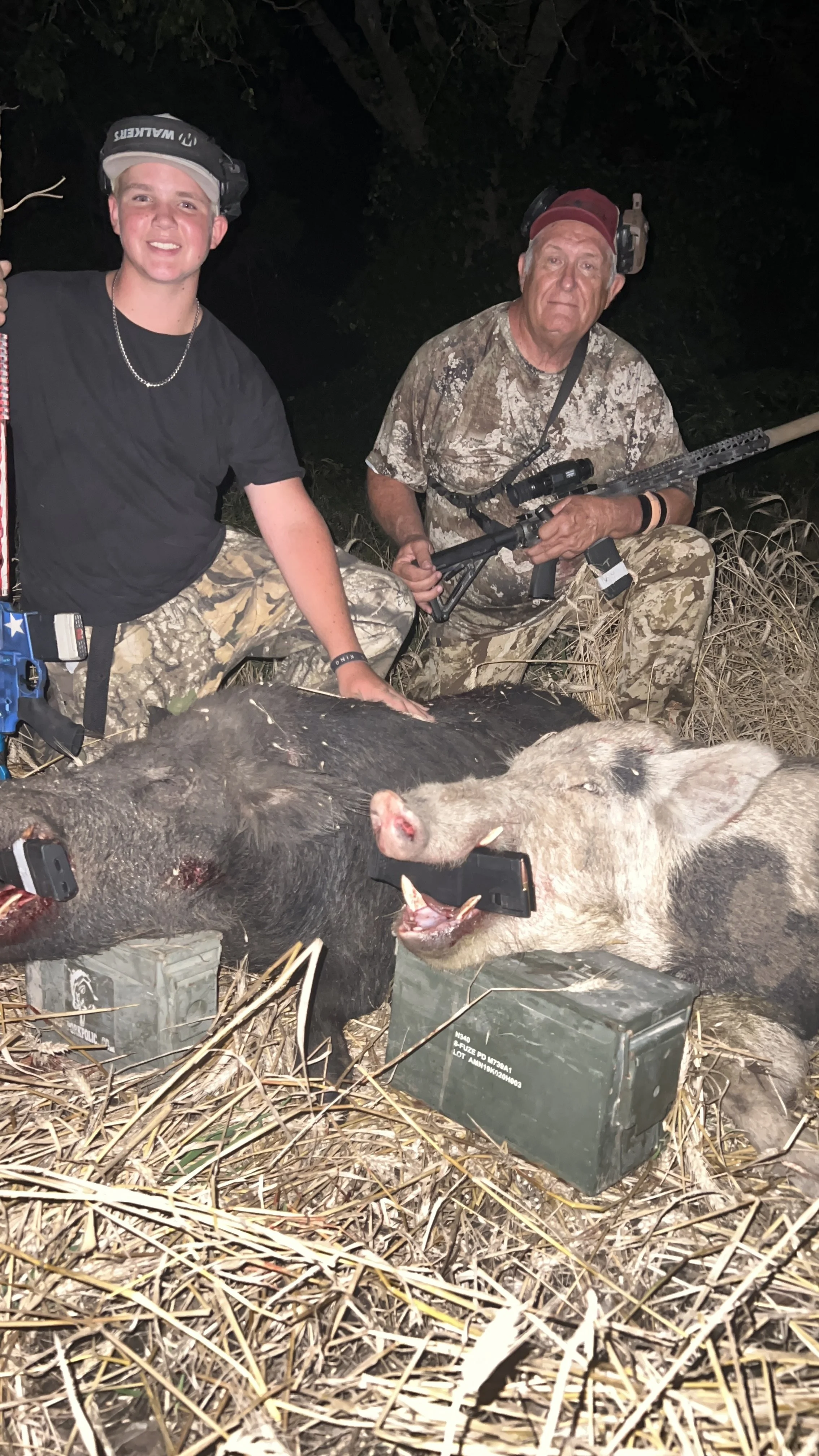 Two men in camouflage clothing and headlamps posing with dead wild pigs at night, with hunting equipment and ammunition nearby, in a natural outdoor setting.