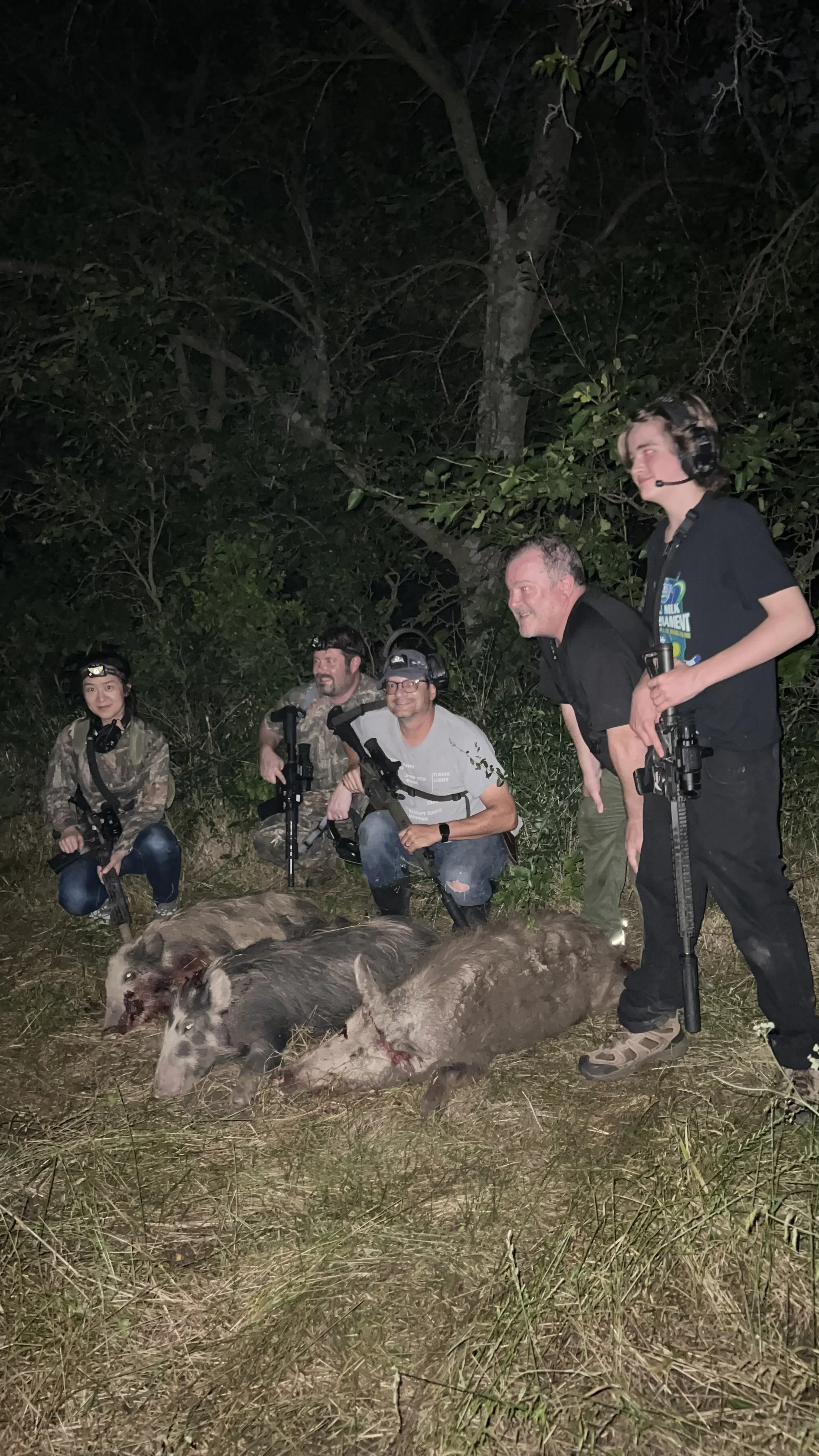 A group of five people kneeling and standing behind two hunting dogs with carcasses lying on the ground, in a wooded outdoor setting at night.