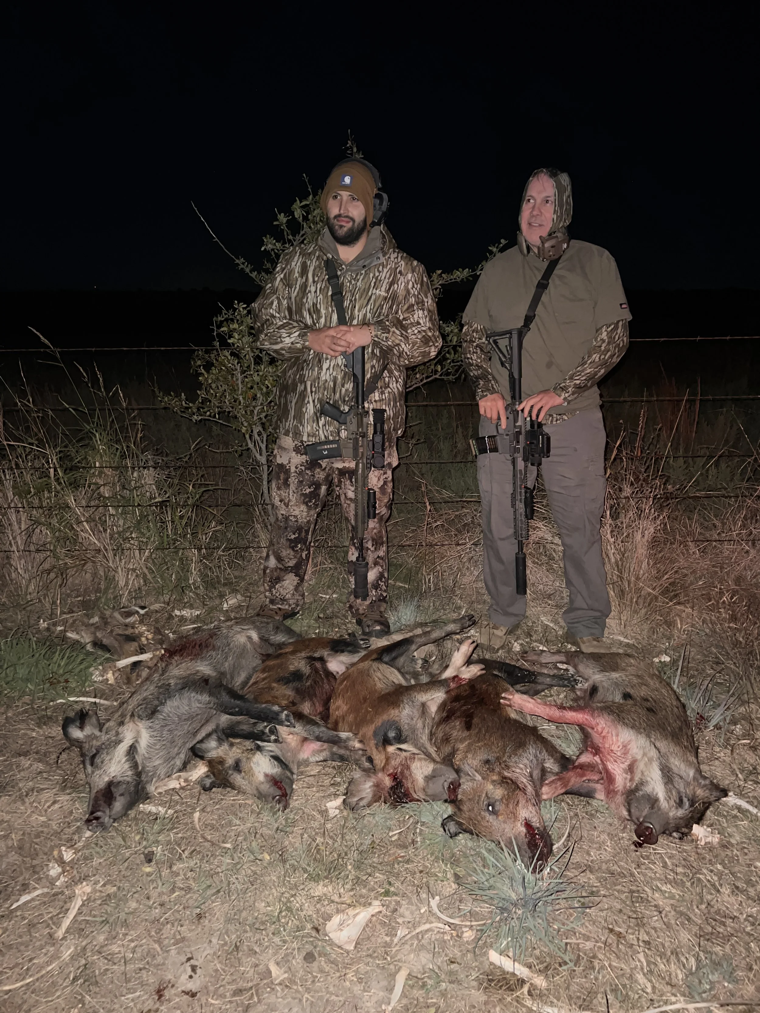 Two men in camouflage and outdoor gear stand behind a fence at night, with multiple dead animals and blood on the ground in front of them.