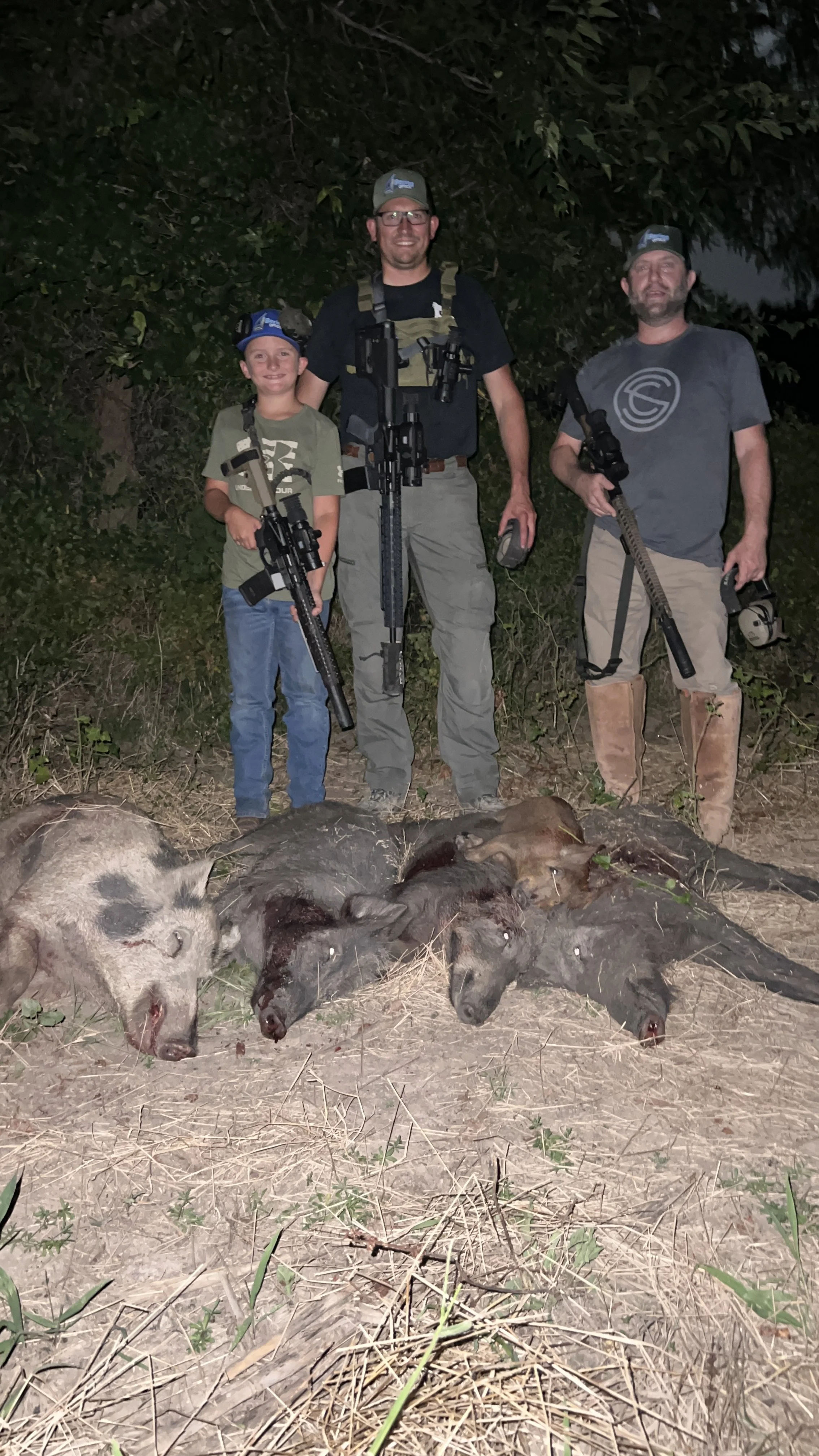 Three hunters are standing behind the carcasses of five pigs in a wooded area at night. They are holding rifles and smiling.
