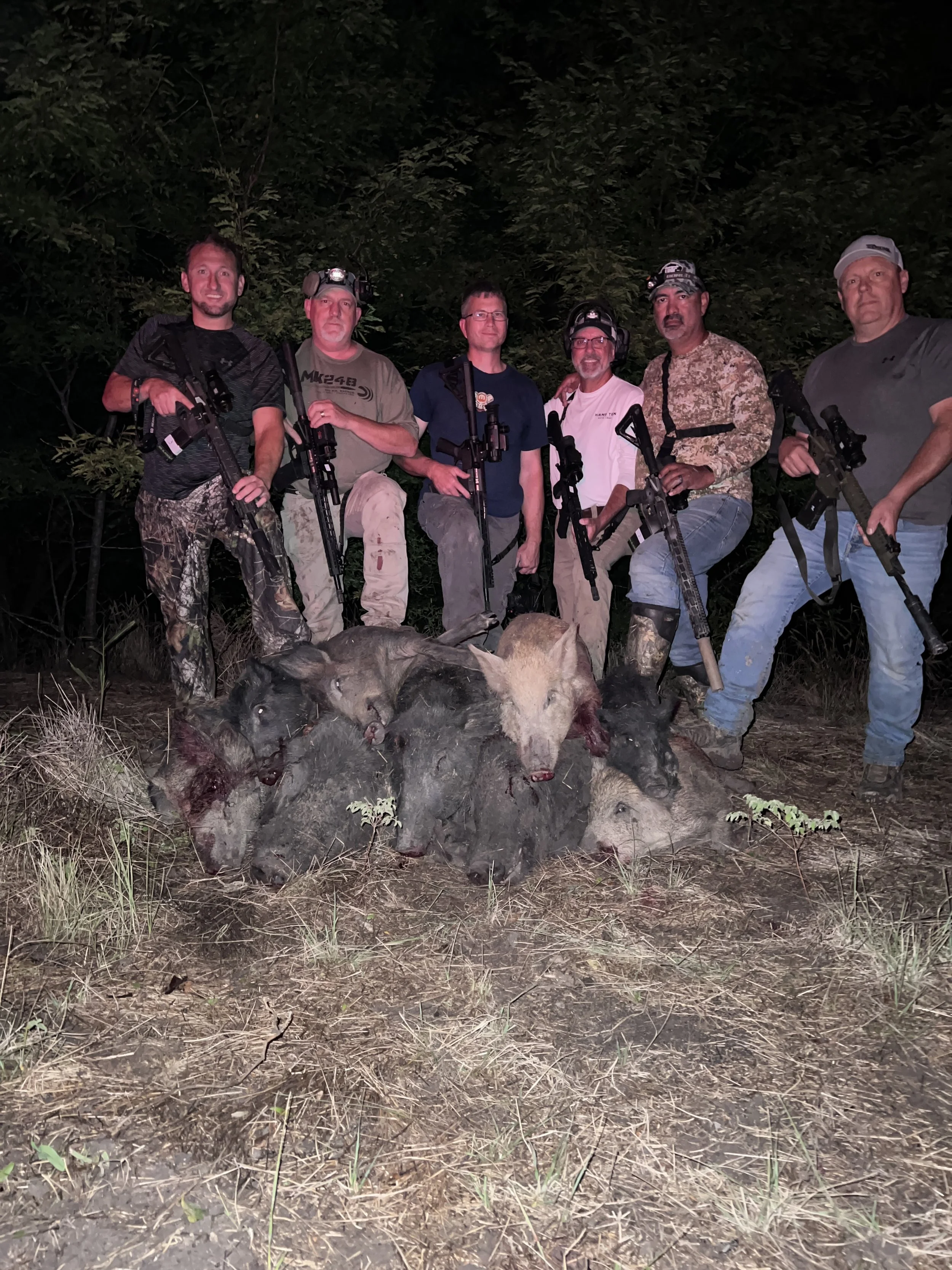 A group of six men posing in a forest at night, with hunted wild animals, including boars, lying on the ground in front of them. All are holding rifles and wearing hunting gear, some with headlamps.