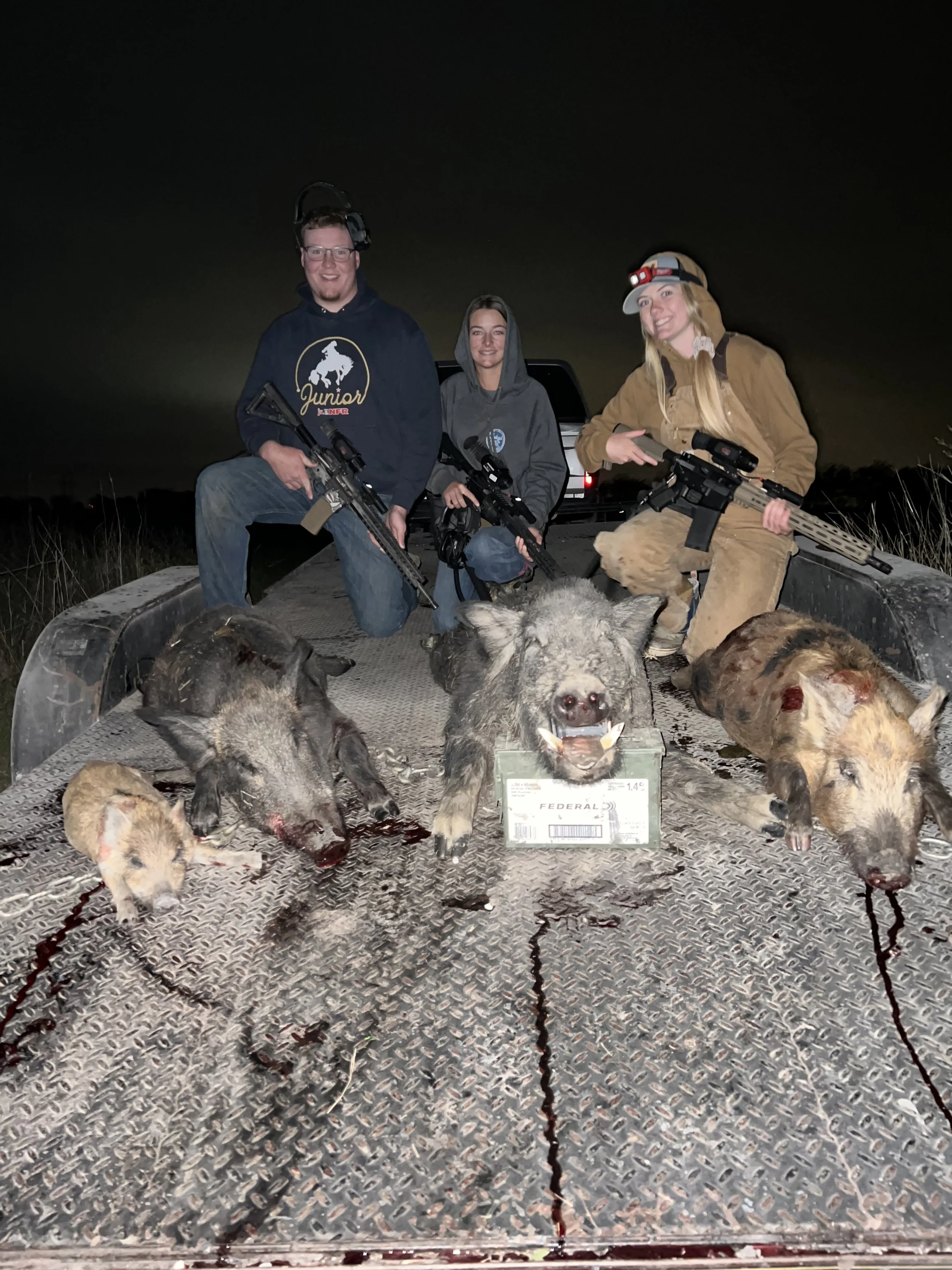 Three people kneeling on a truck bed at night, holding rifles, with slain wild boars and blood on the metal surface in front of them.