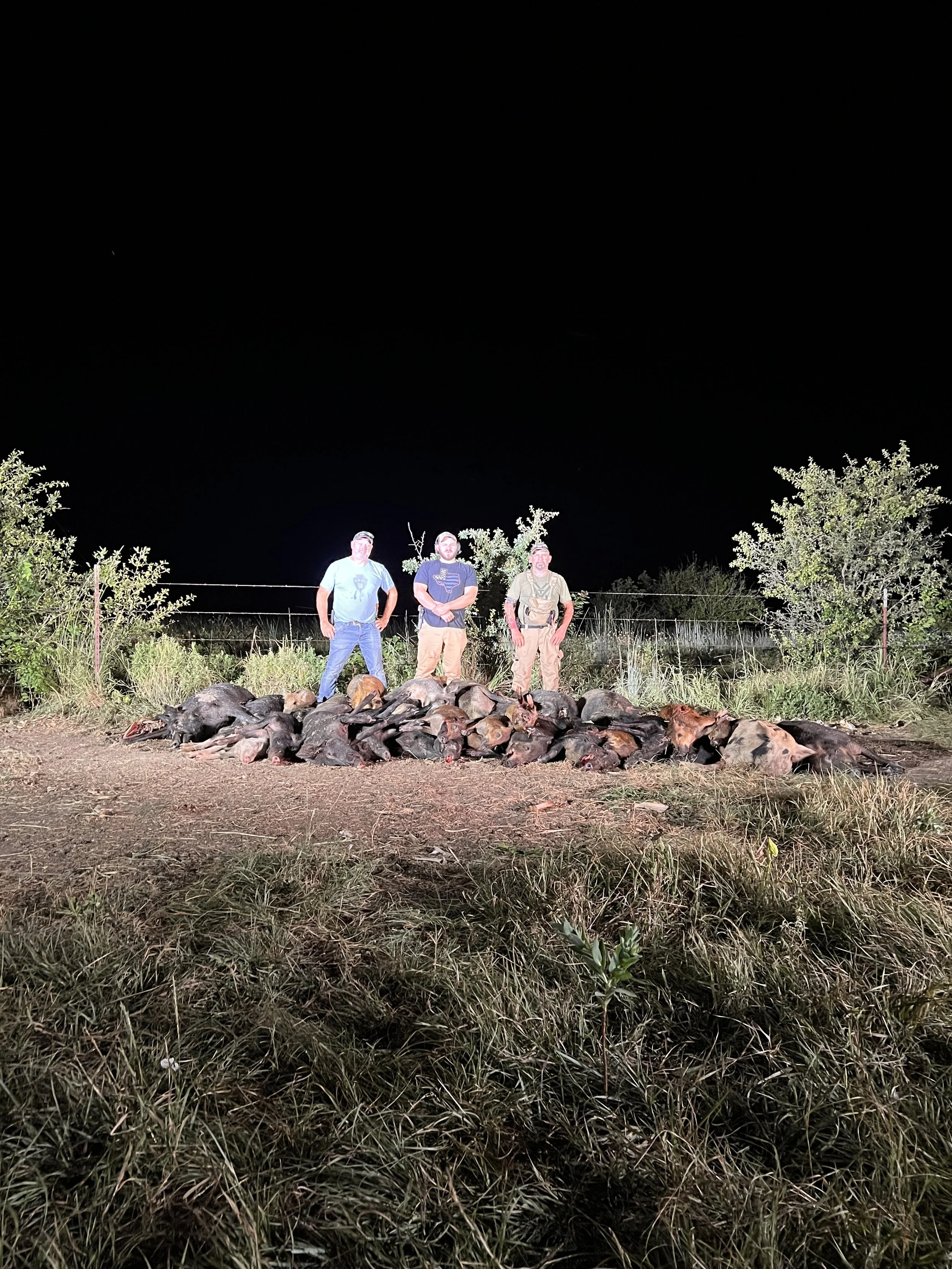 Three men stand outdoors at night behind a pile of animal carcasses in a field with bushes and trees, illuminated by a bright flashlight or camera flash.