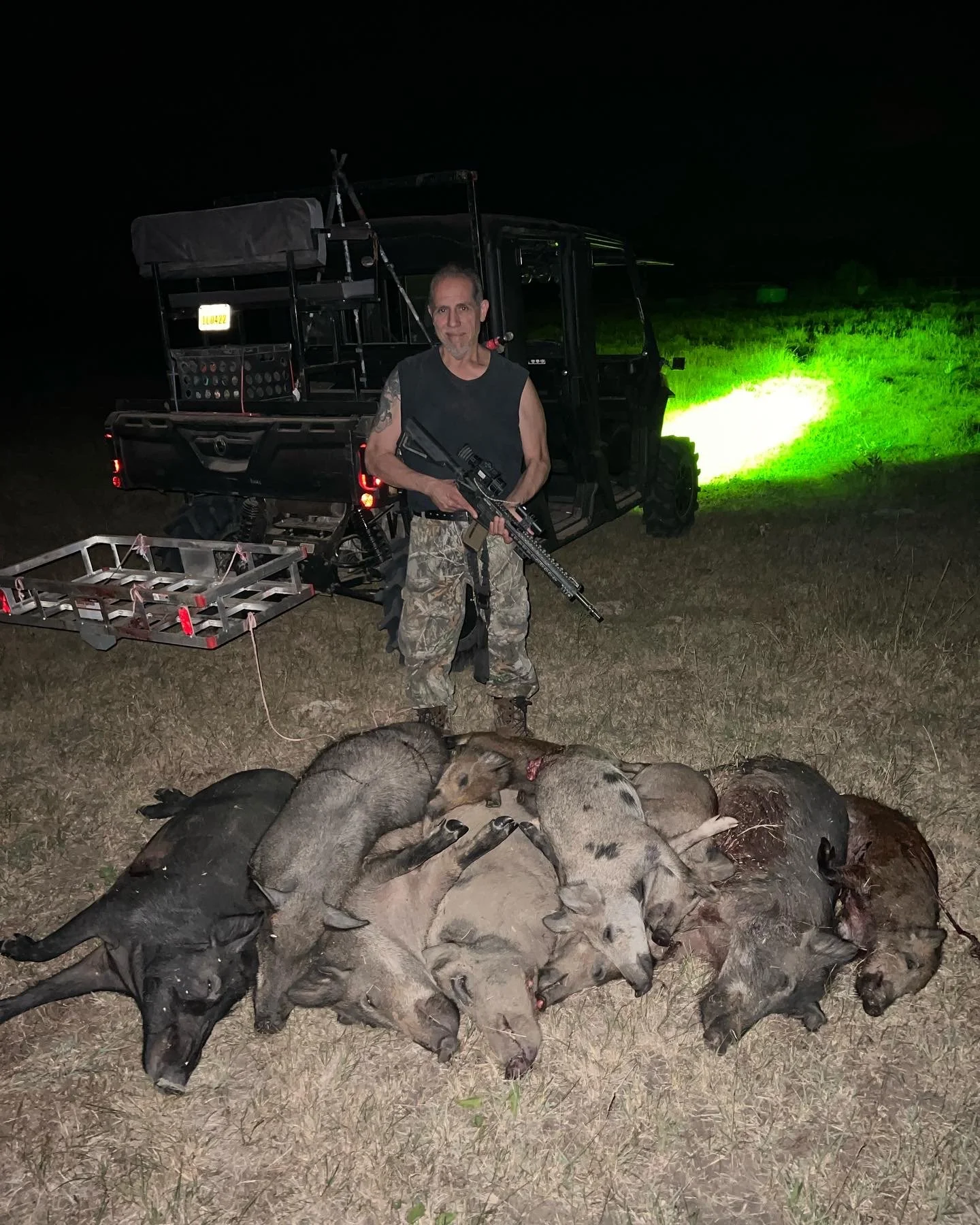 A man standing outdoors at night, holding a rifle, with a utility vehicle behind him, a pile of hunted boars and pigs in front, and a green light shining in the background.