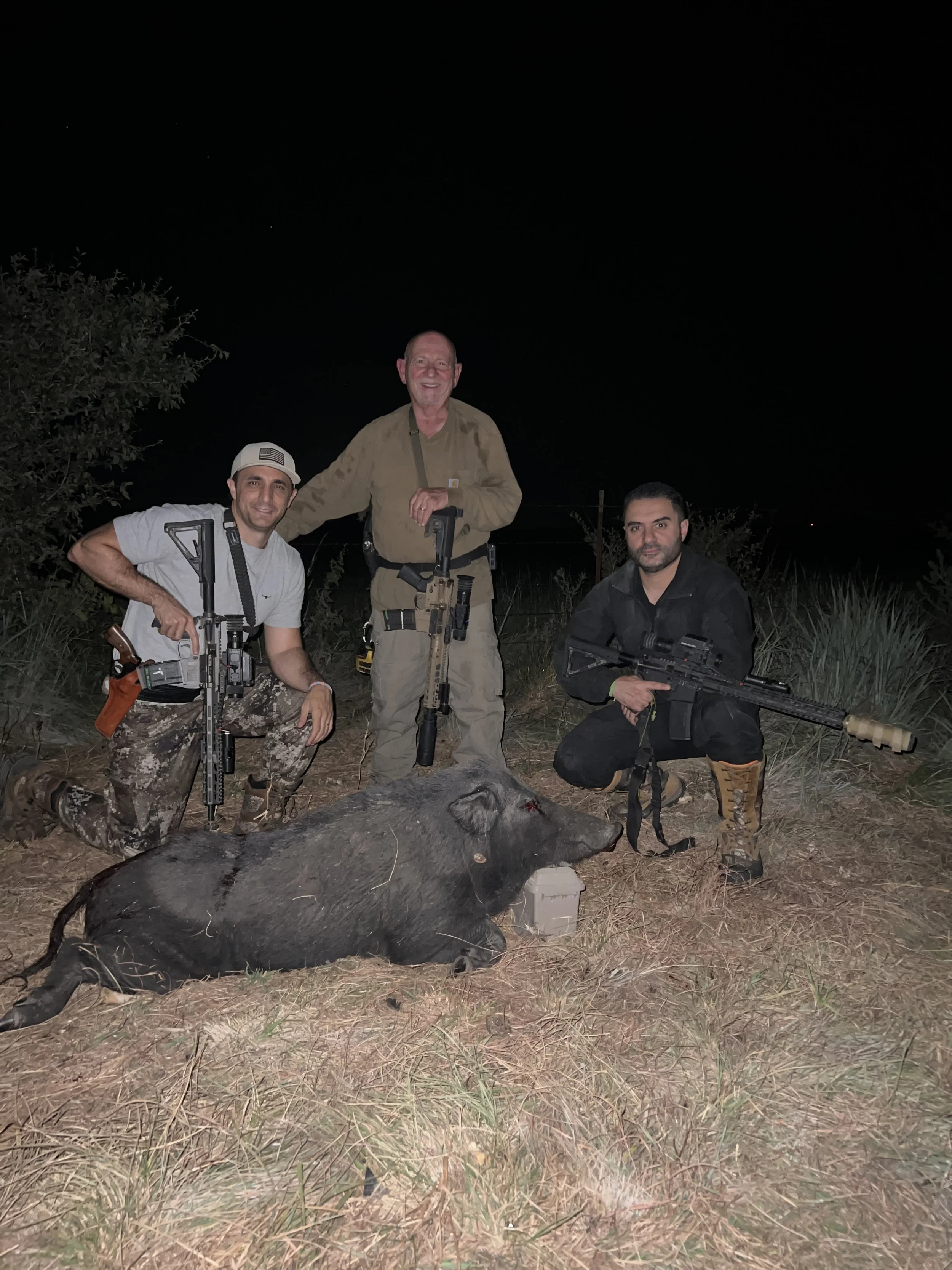 Three men with firearms and tactical gear kneel and stand around a freshly killed animal on the ground at night.