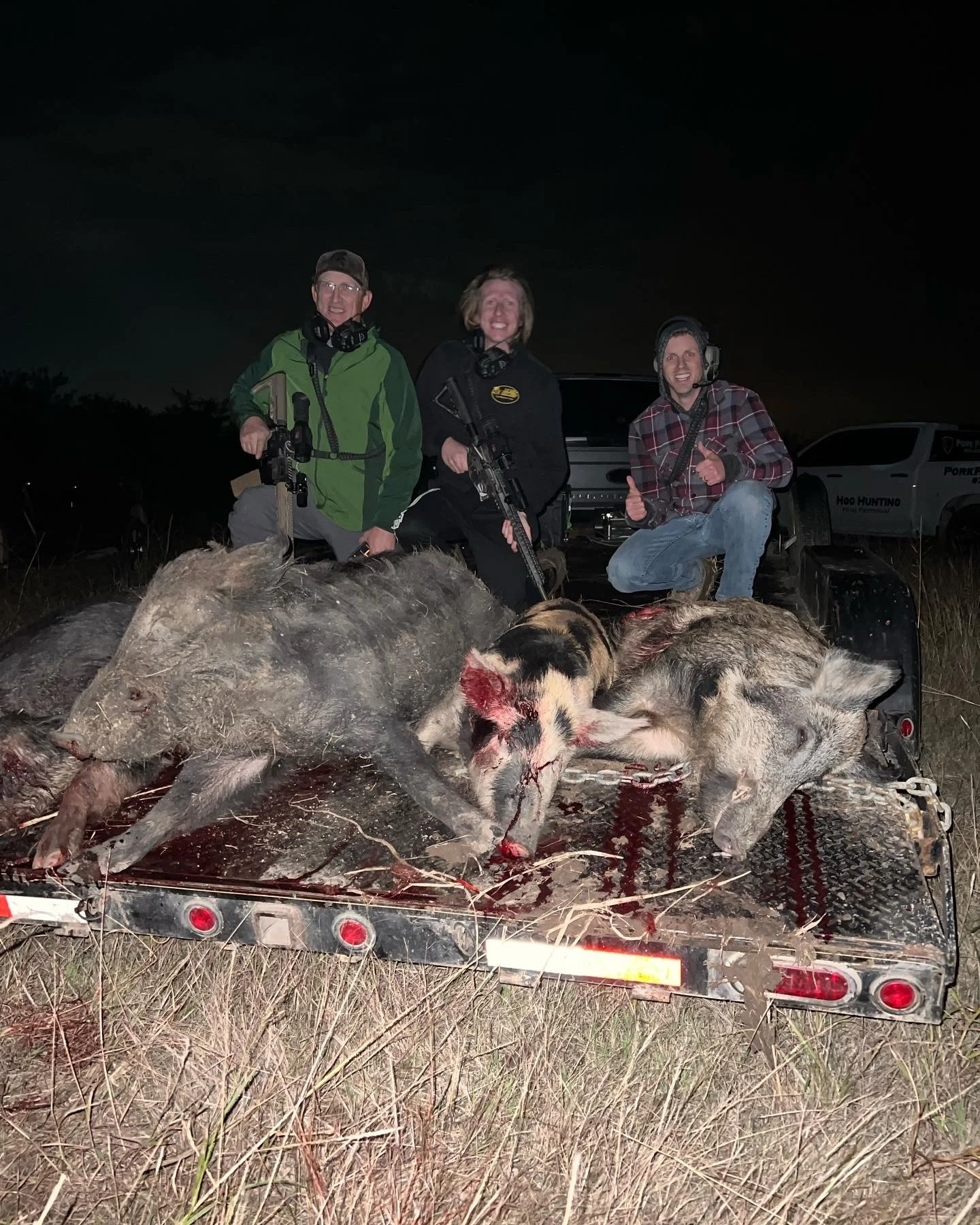 Three people smiling and holding guns on a truck bed, with two dead pigs covered in blood in front of them, at night outdoors.