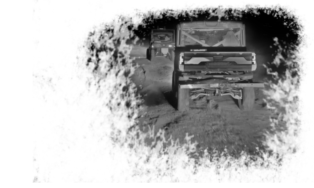 Nighttime view of three off-road vehicles driving on a rugged desert terrain, with the front vehicle's headlights illuminating the path.