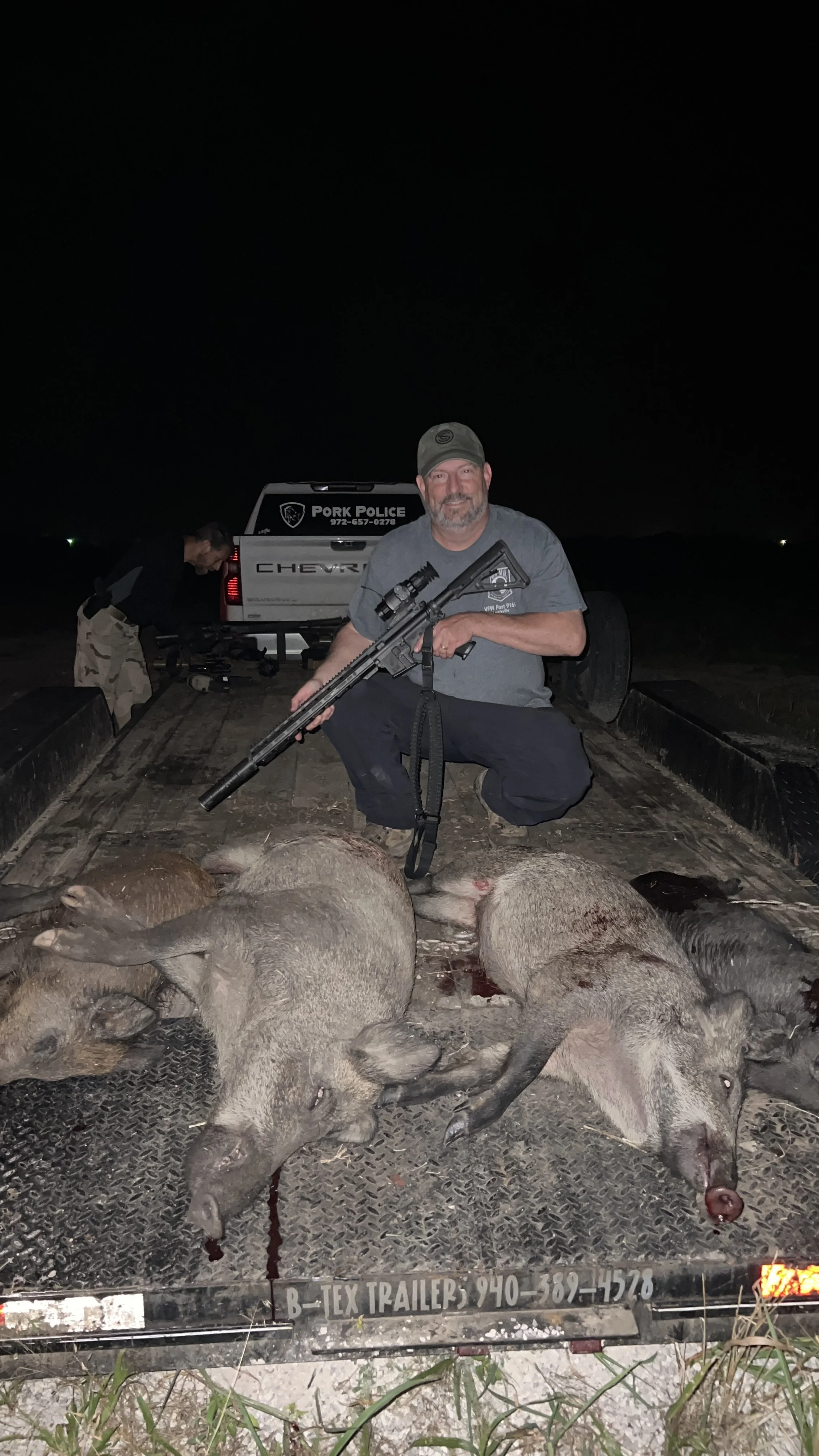 A man squatting on a trailer with a rifle and six dead pigs laid out in front of him, with another person in the background near the truck.