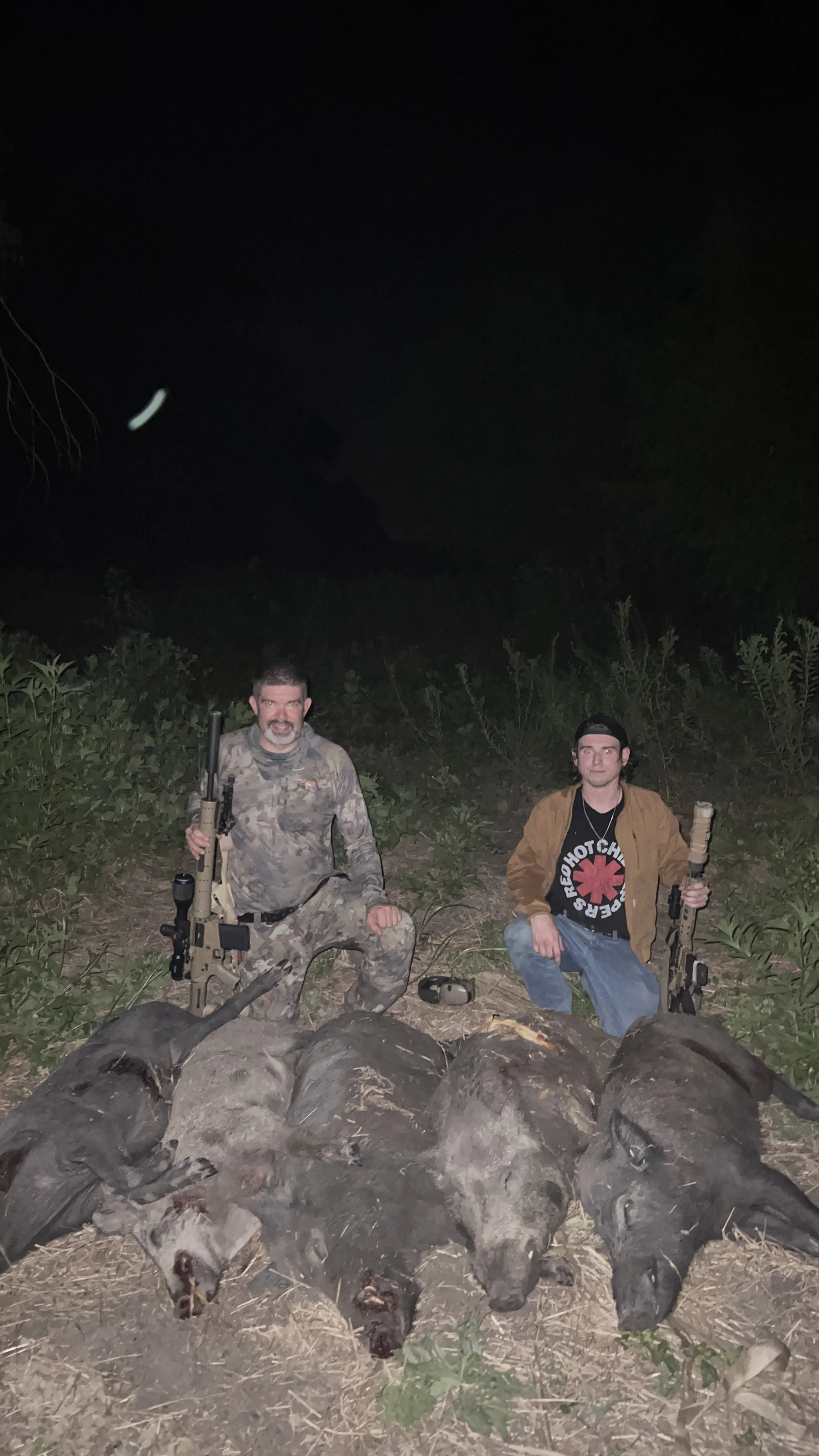 Two men kneeling on the ground at night, surrounded by large animals laid on their backs. Both men hold rifles, and are wearing casual clothing; one in camouflage and the other in a brown shirt with a Red Cross logo. The background is dark with some 
